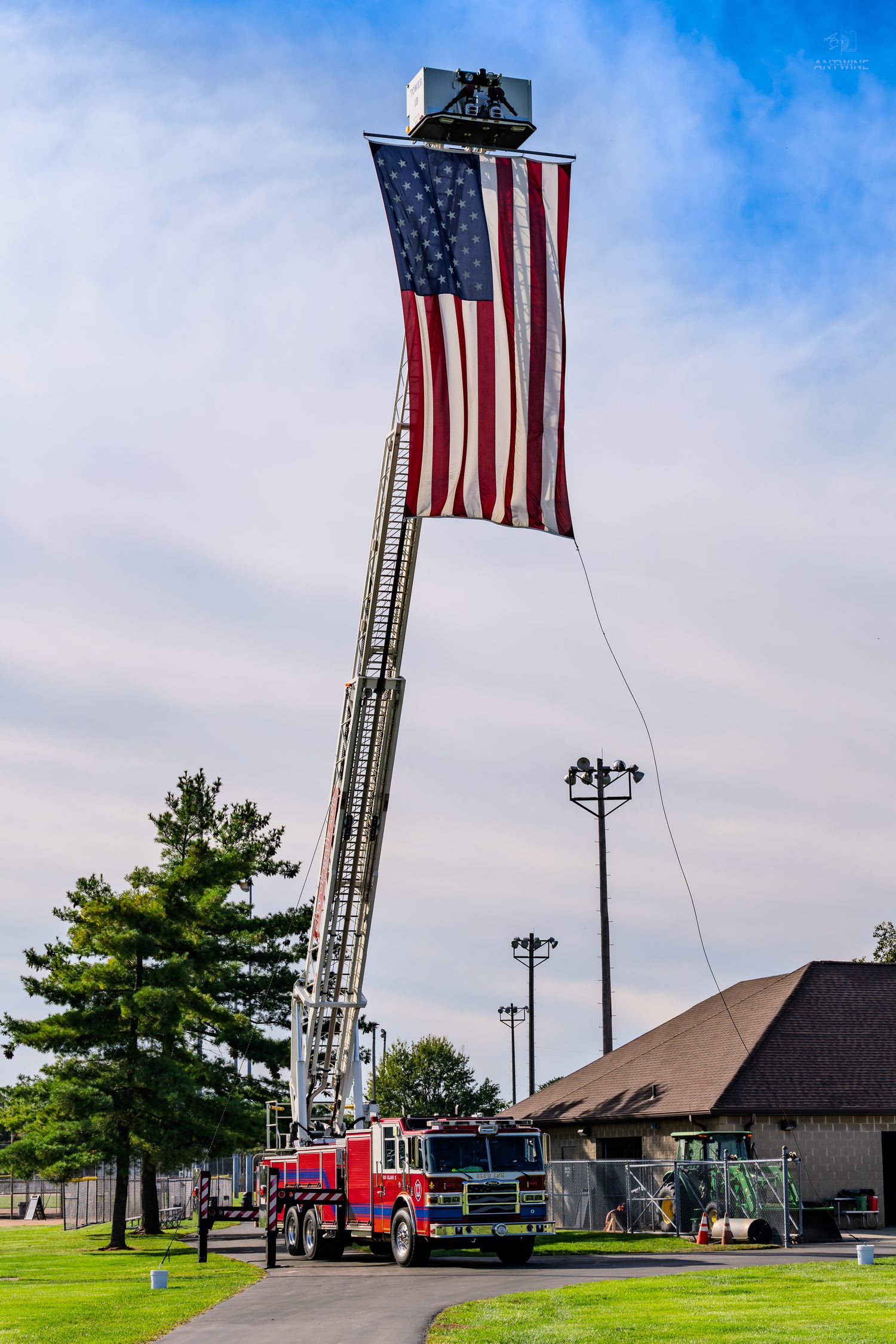 Red fire truck with extended ladder displaying large American flag against blue sky.