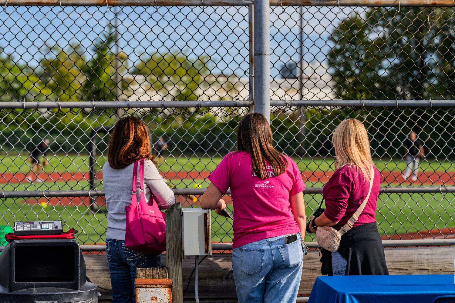 Three women in pink shirts watch a sports field through a chain-link fence on a sunny day.