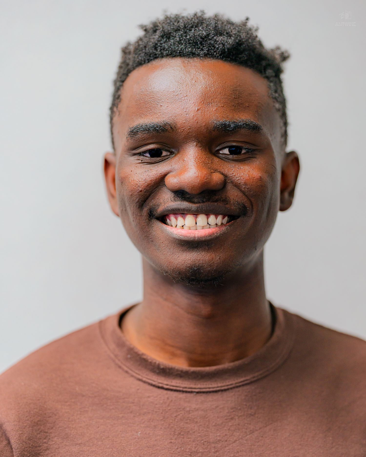 Smiling young Black man with short hair wearing a brown sweatshirt against a light gray background.
