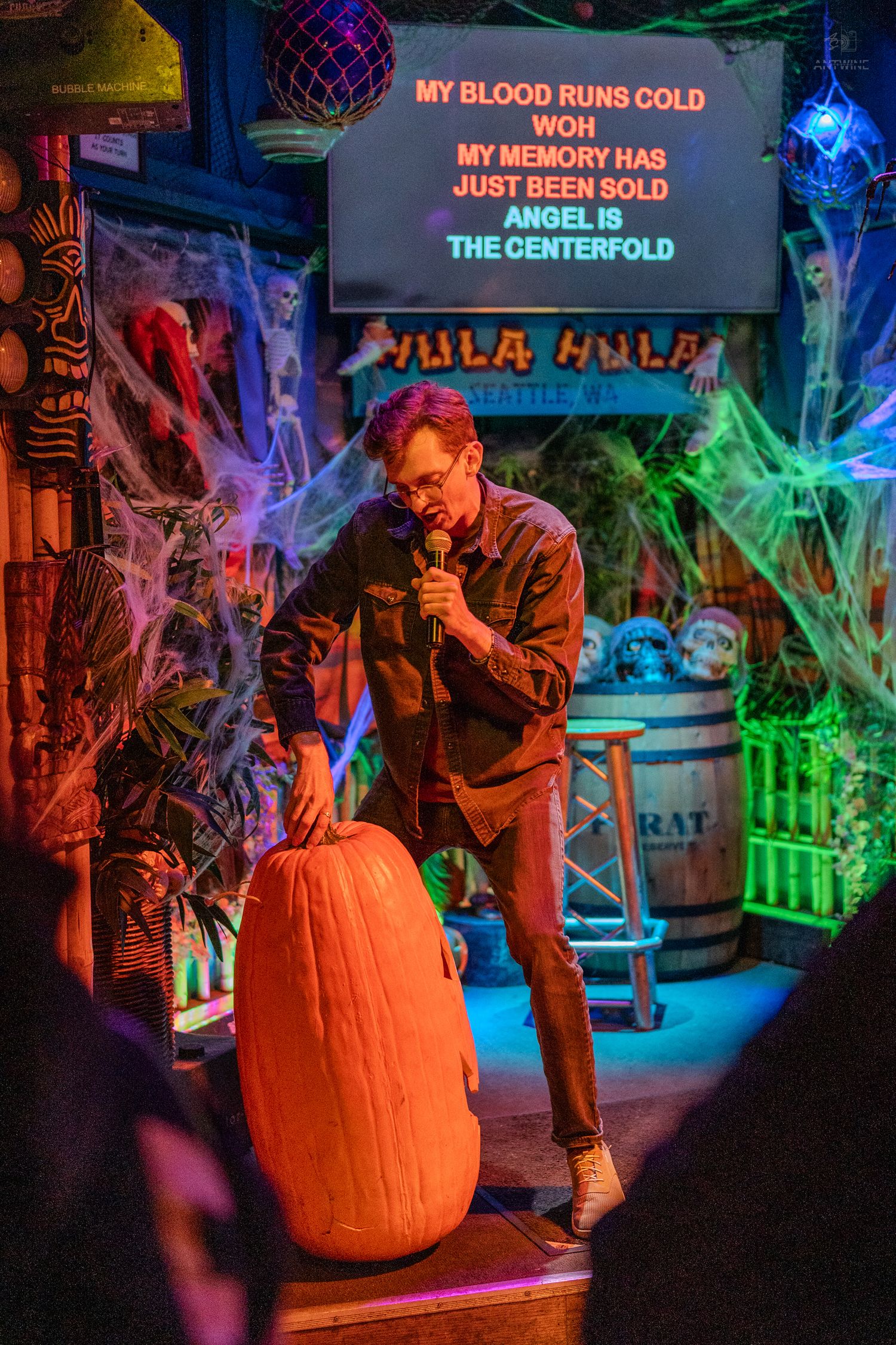 Man singing karaoke on Halloween-decorated stage at Hula Hula bar in Seattle, holding microphone near large pumpkin.