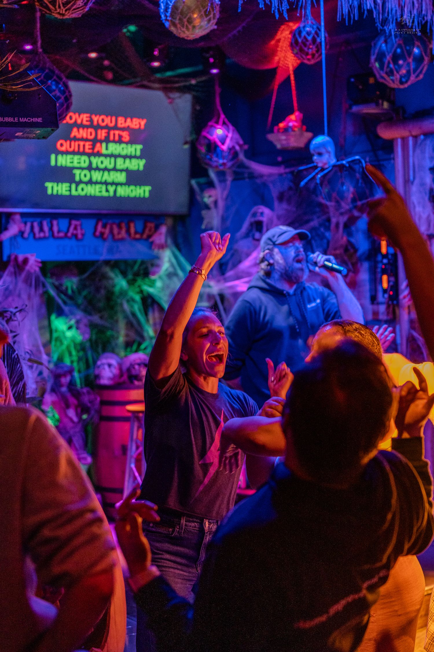 Excited crowd dancing at Halloween karaoke night in neon-lit Hula Hula bar with festive decor.
