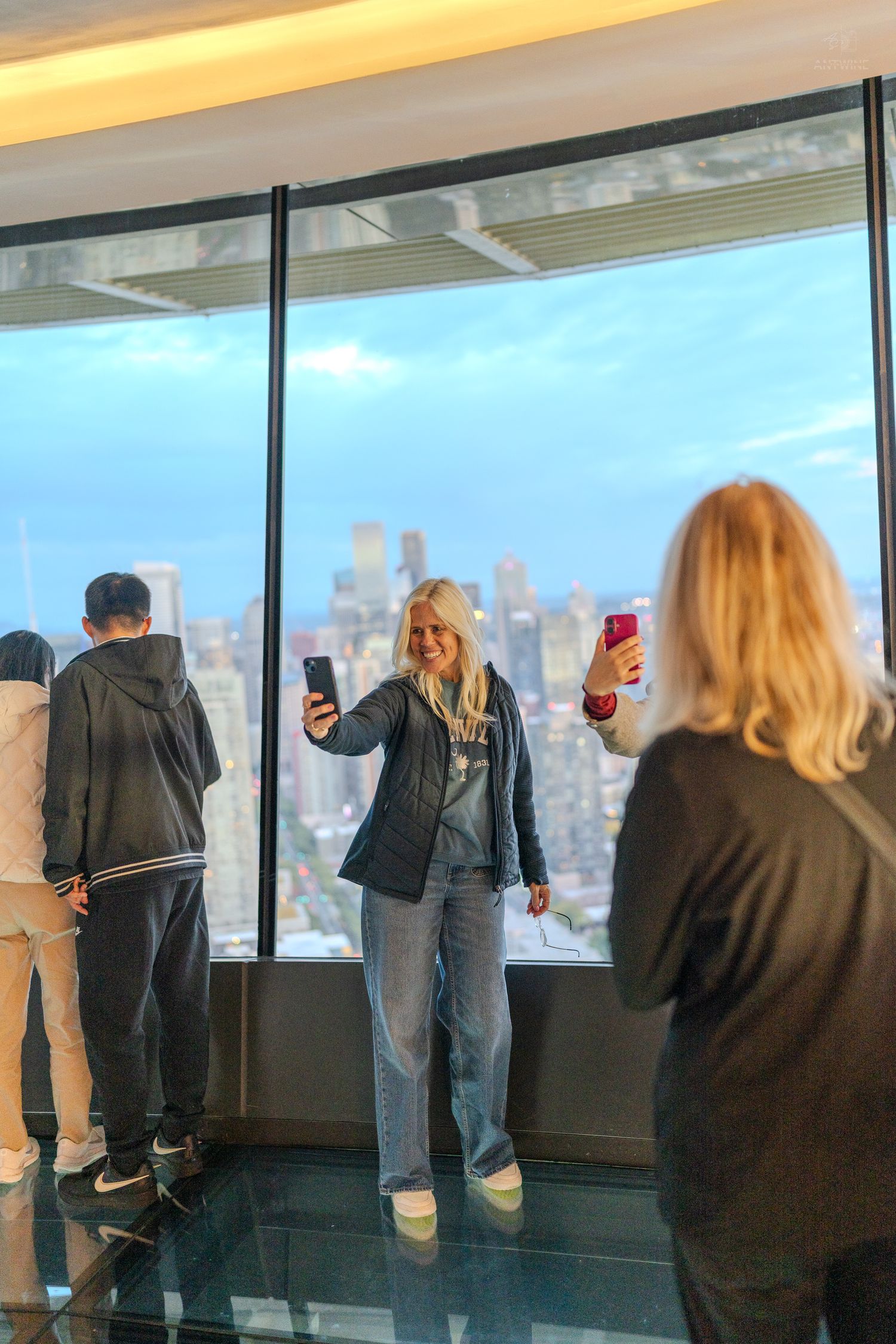 A blonde woman takes a selfie at a skyscraper observation deck with a city skyline view.