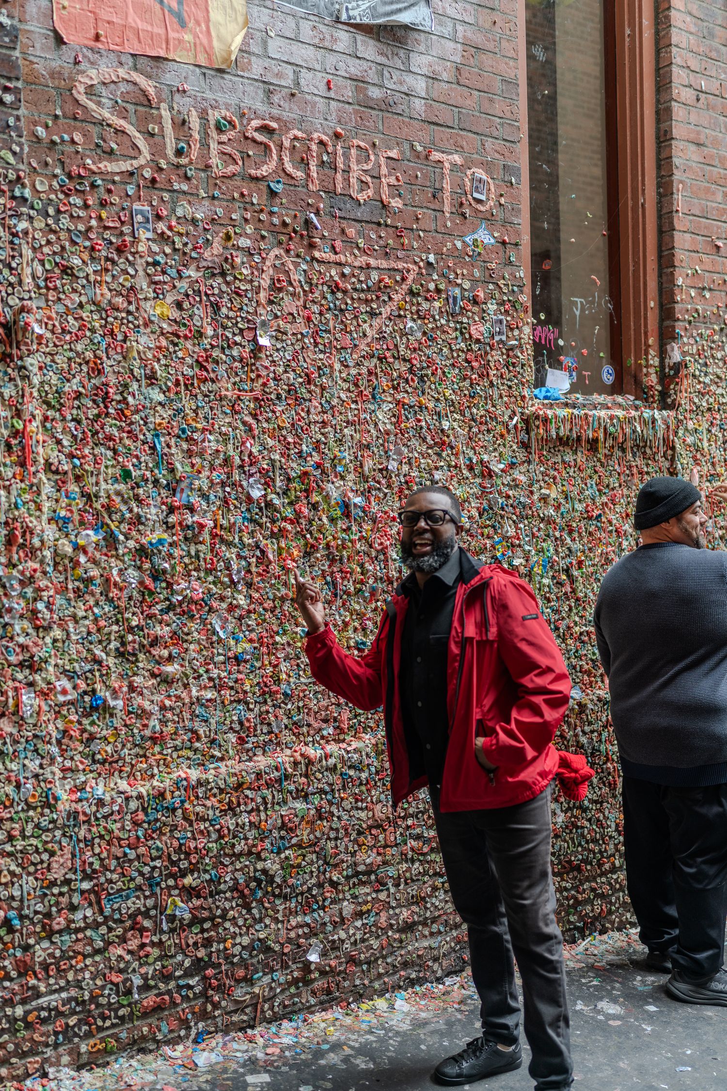 Man in red jacket poses at Seattle's famous Gum Wall, covered in colorful chewing gum with Subscribe To written on it.