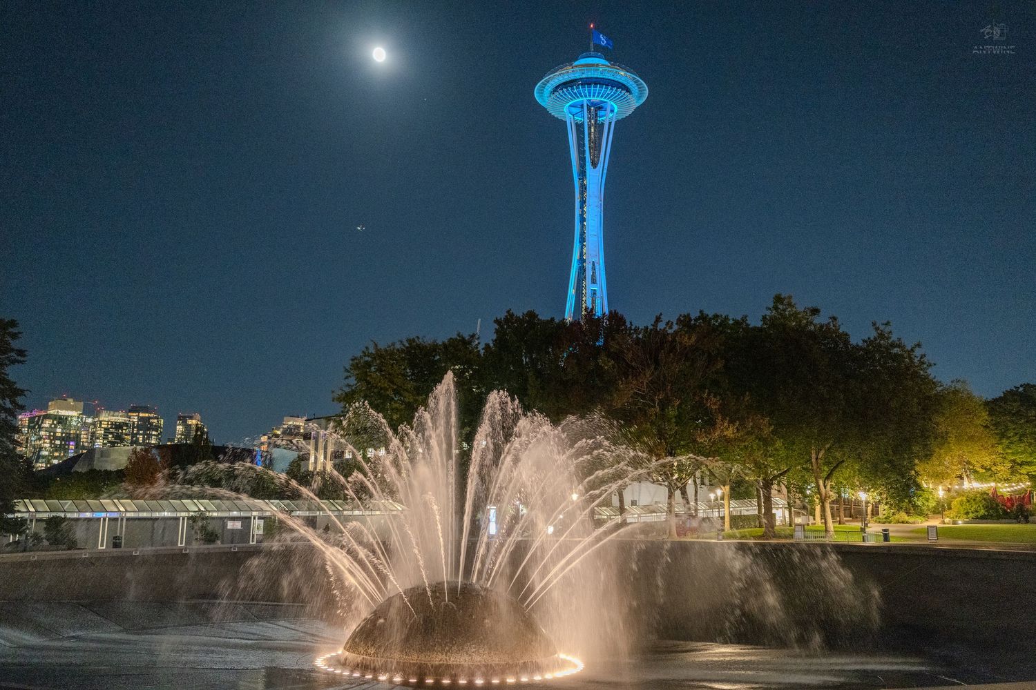 Seattle Space Needle lit in blue at night with a fountain and full moon in the background.