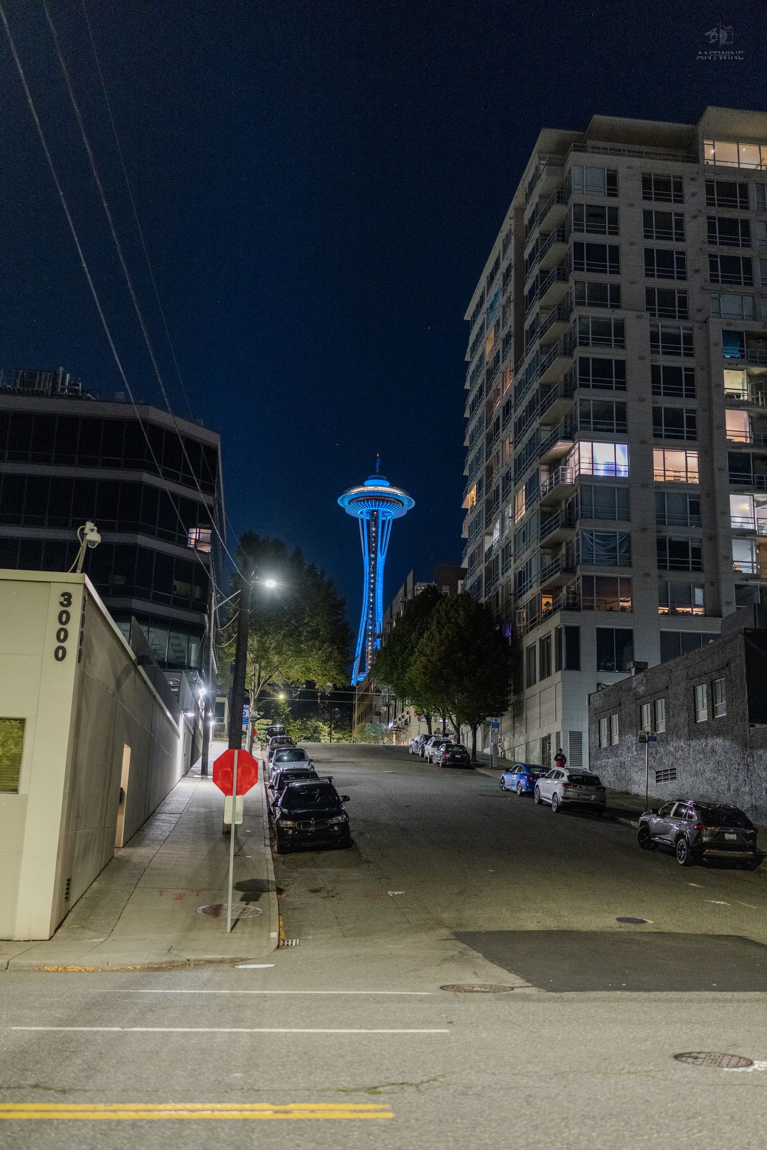 Seattle Space Needle illuminated in blue light at night, viewed from a quiet urban street flanked by modern buildings.