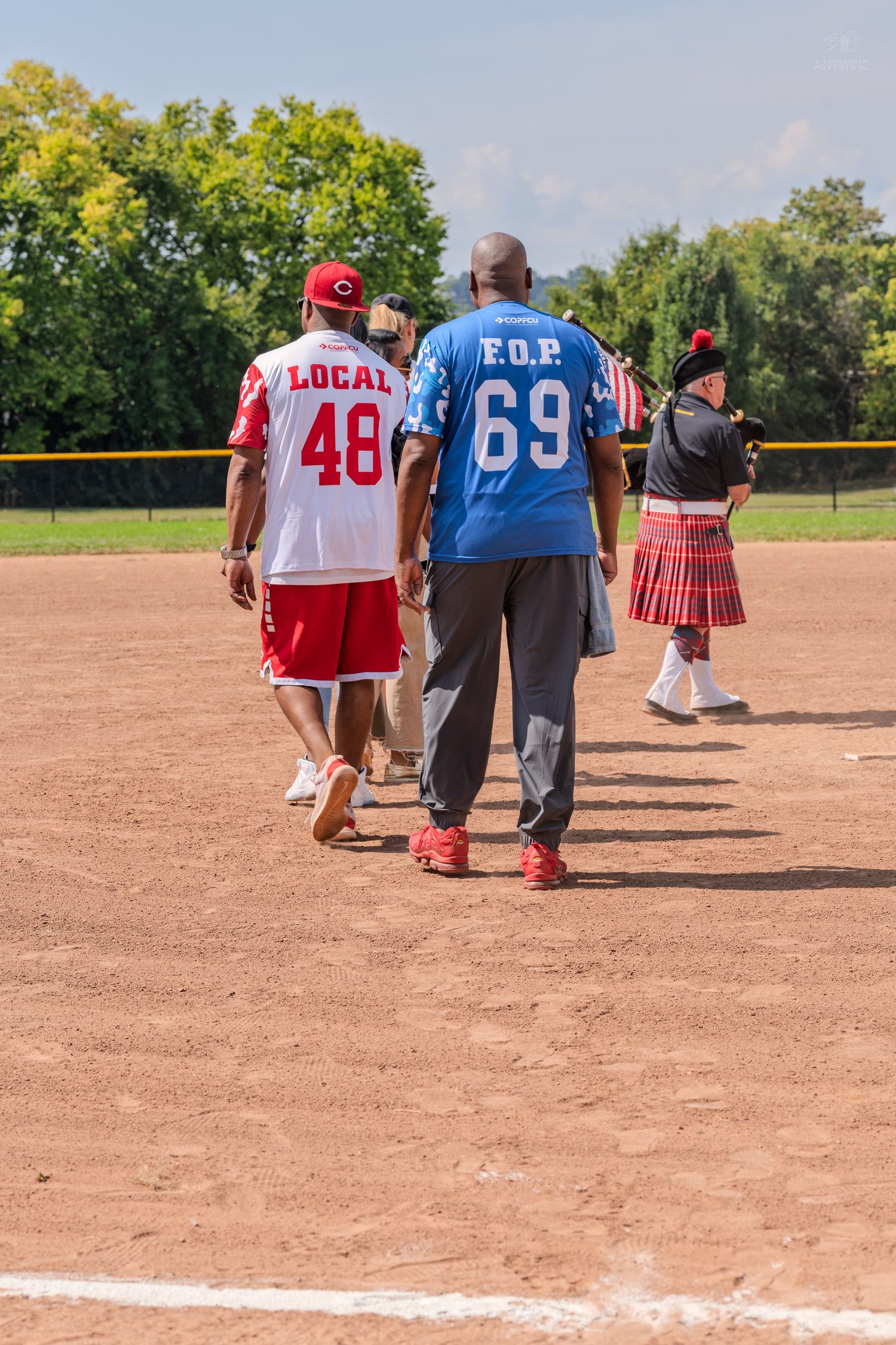 Two men in sports jerseys walk across a baseball diamond, with a man in a kilt visible in the background.