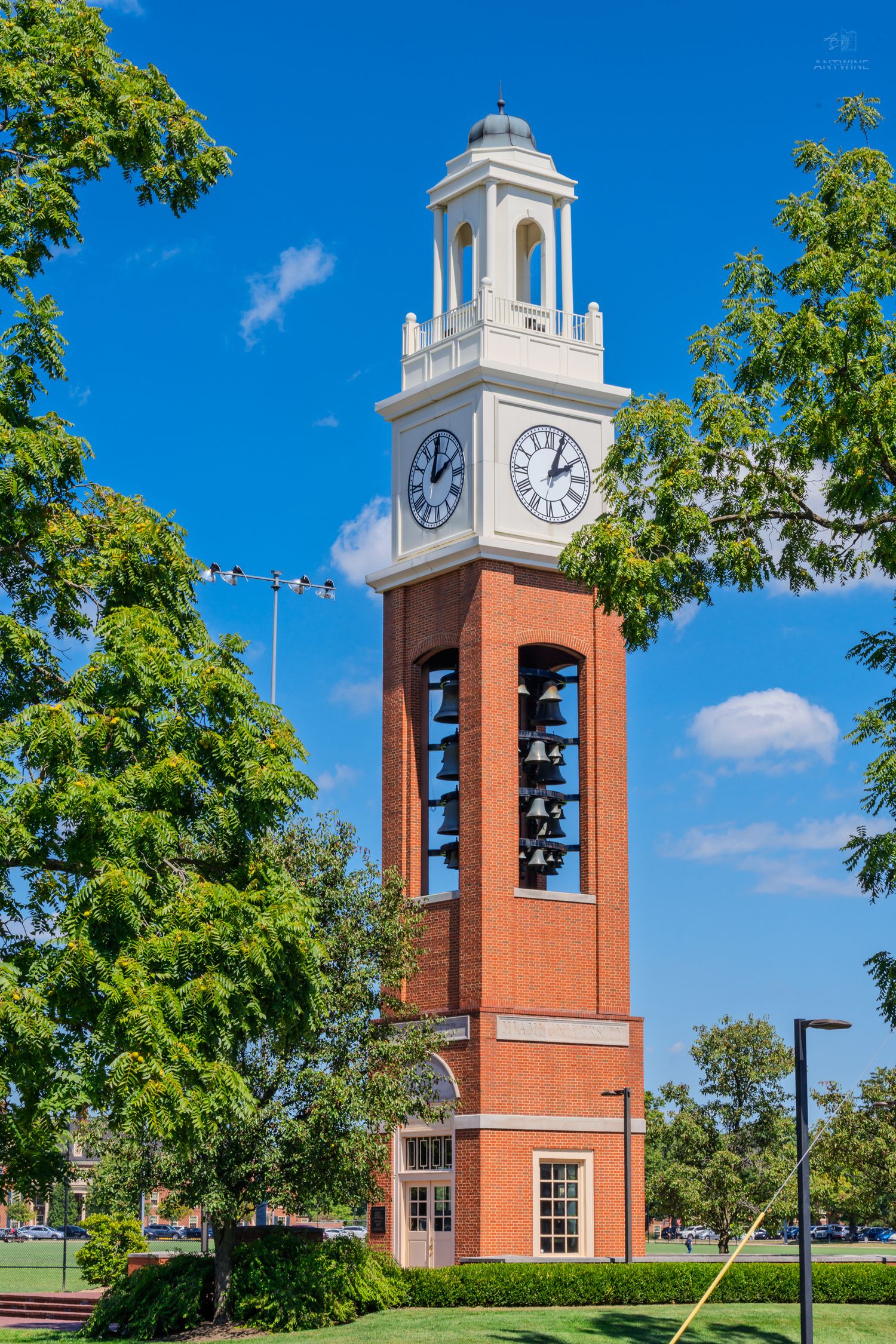 Historic red brick carillon clock tower surrounded by lush green trees under a bright blue sky.