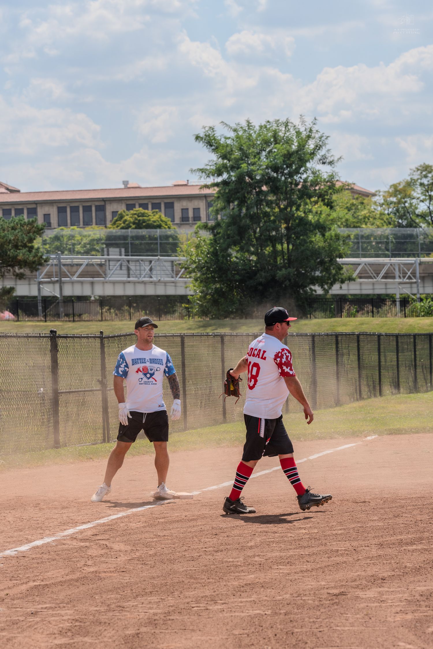Two softball players on a dirt infield during a recreational game on a sunny day with blue skies.