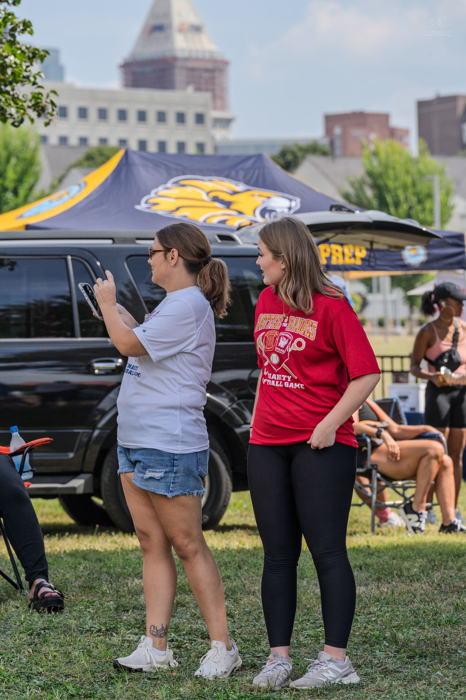 Two women standing on grass at an outdoor tailgate event with tents and a van in background.