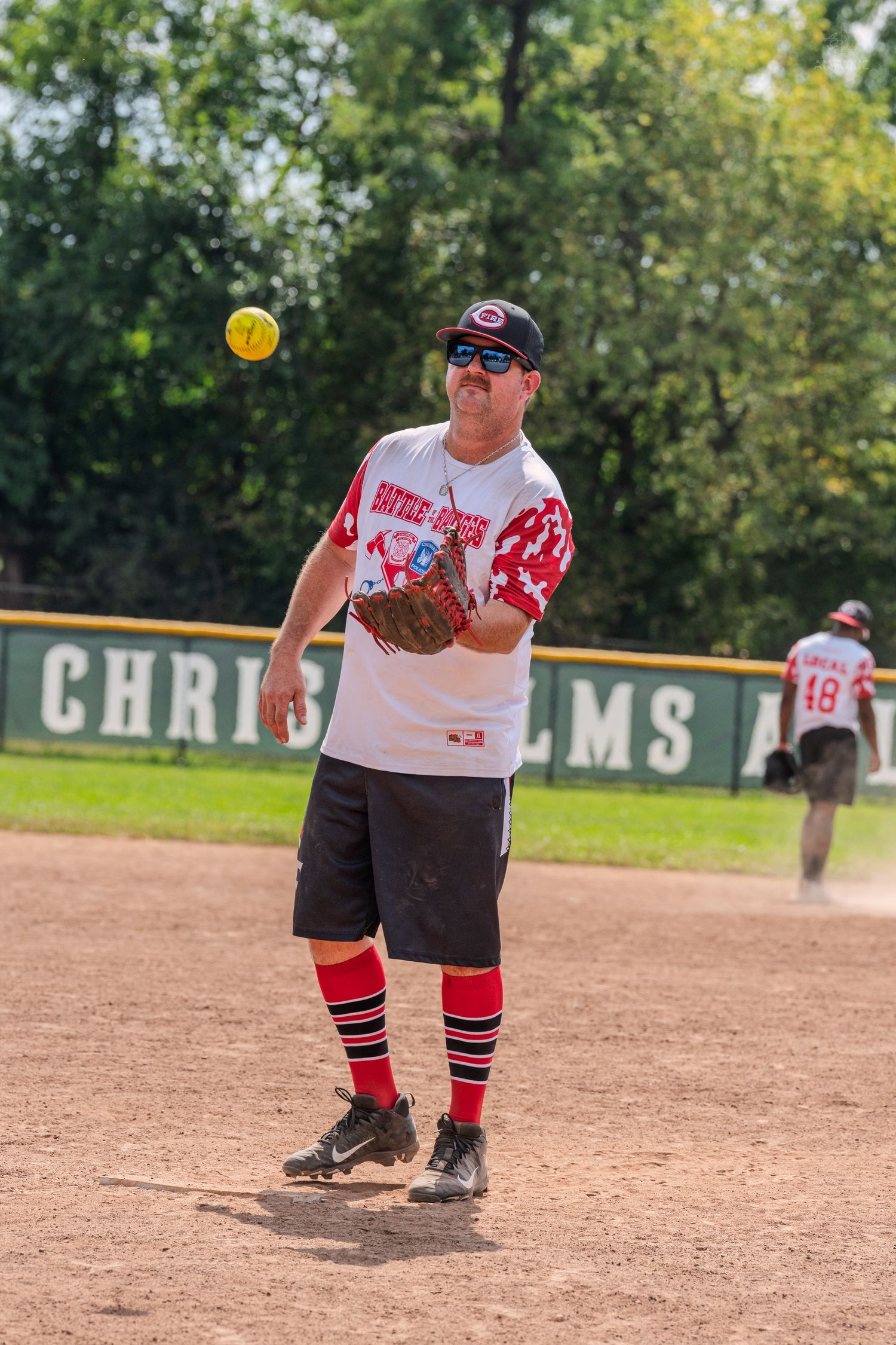 Male softball player throws yellow ball wearing red socks and sunglasses on dirt infield.