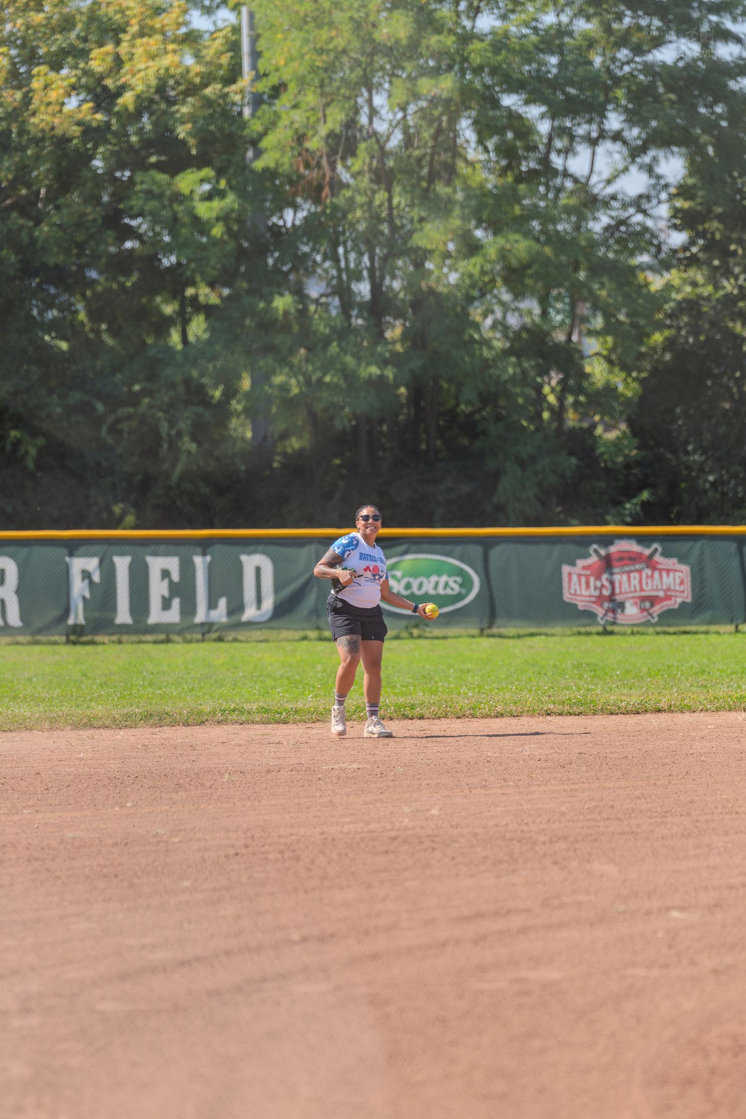 Female softball player in blue jersey preparing to throw ball on dirt infield during All-Star Game at outdoor baseball field.
