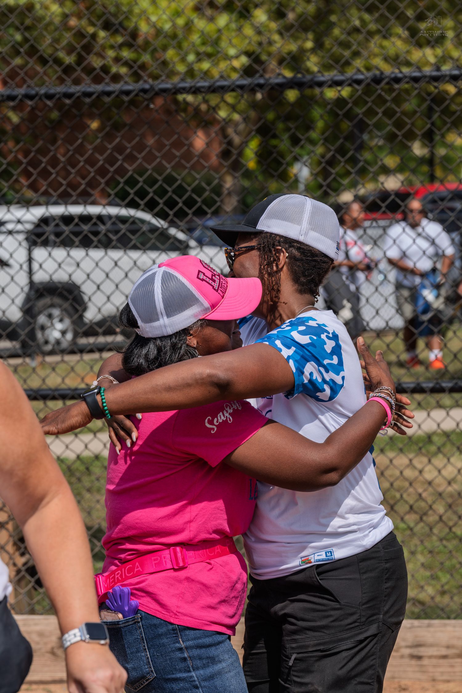 Two women share a warm hug outdoors at a softball event, wearing pink and white jerseys and trucker hats.