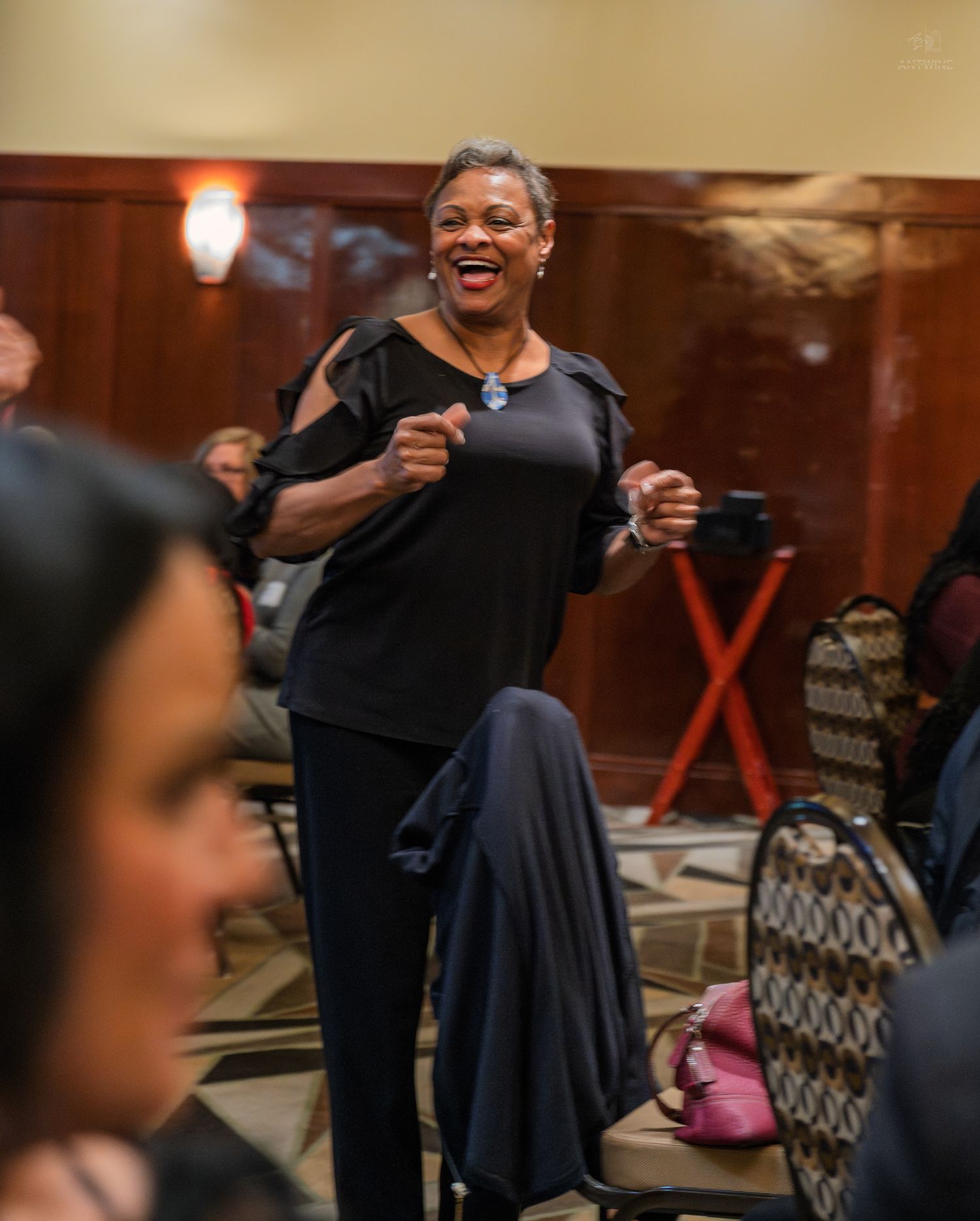 A woman in black smiles and gestures enthusiastically at an indoor networking event.