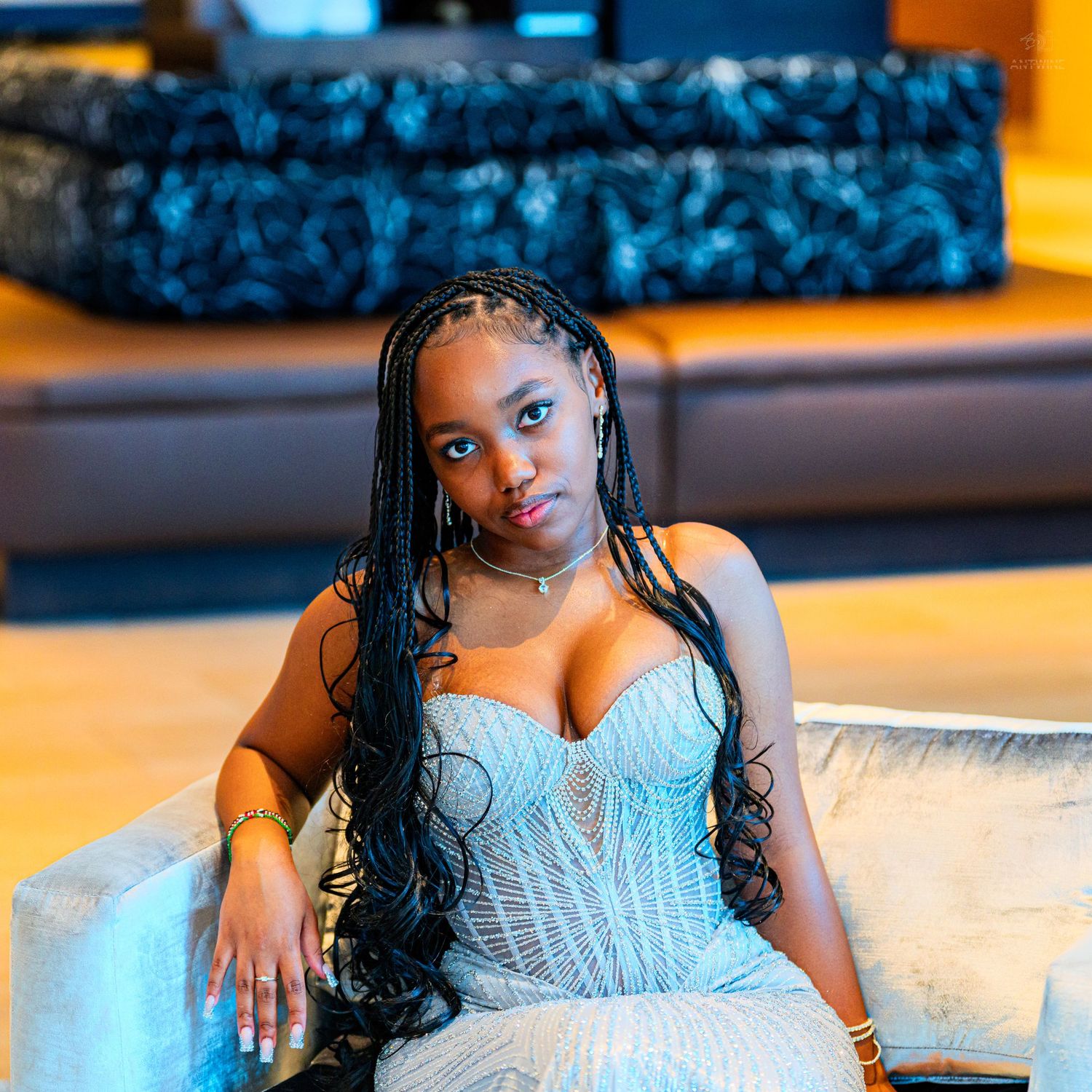 Young woman in a beaded blue strapless gown with long braids seated elegantly in a modern lounge.