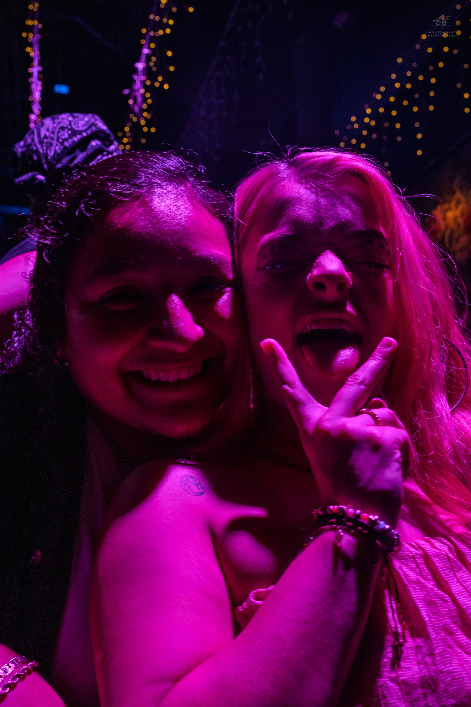 Two smiling women pose together under vivid pink neon lights in a nightclub.