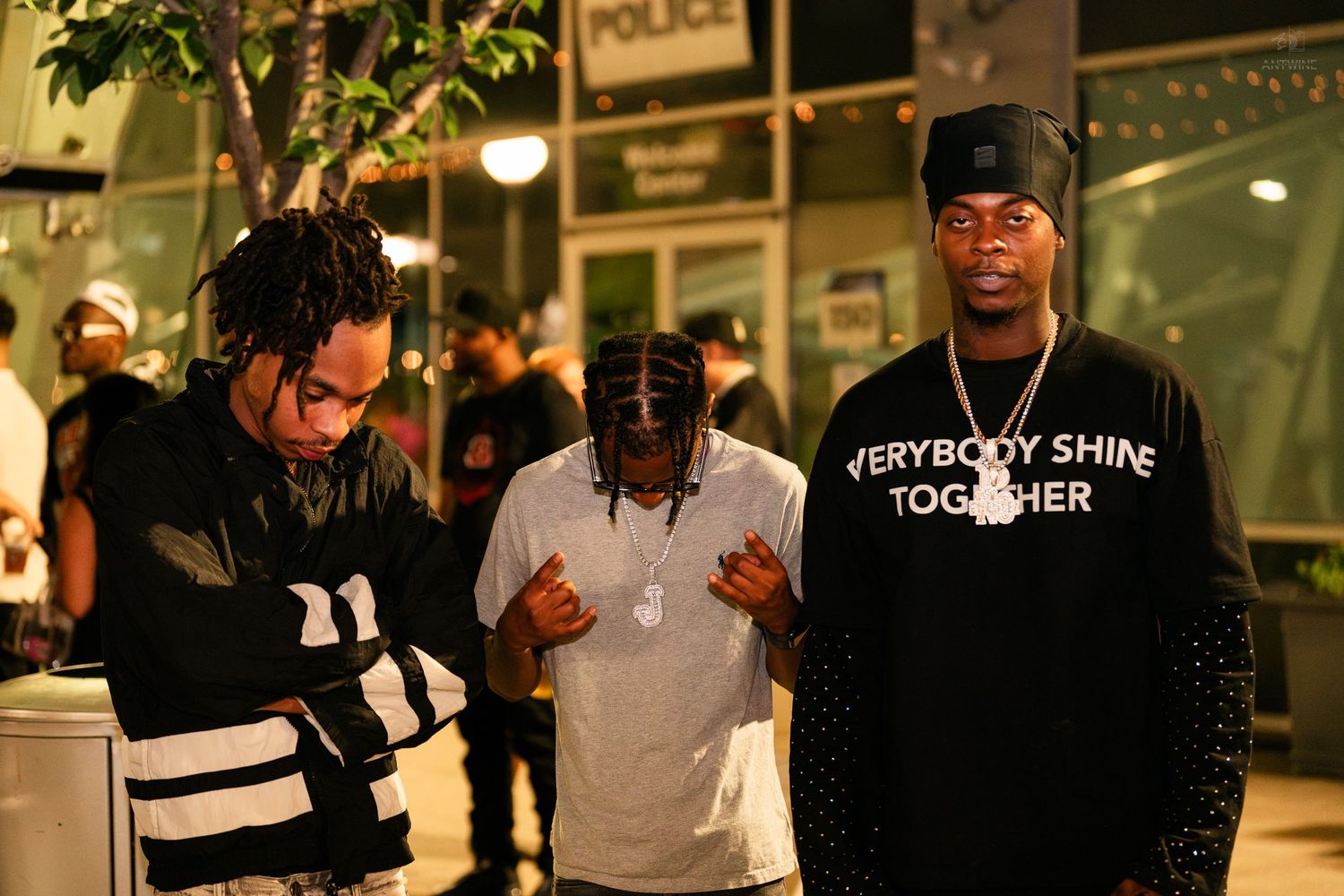 Three men posing outdoors at night, one in Everybody Shine Together black shirt.
