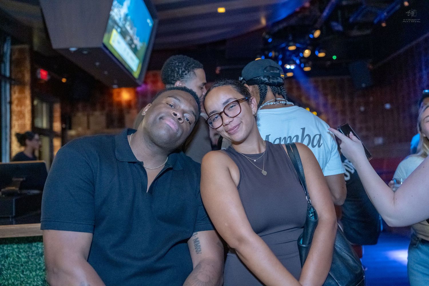 A couple leans together affectionately at a dimly lit bar with blue ambient lighting.