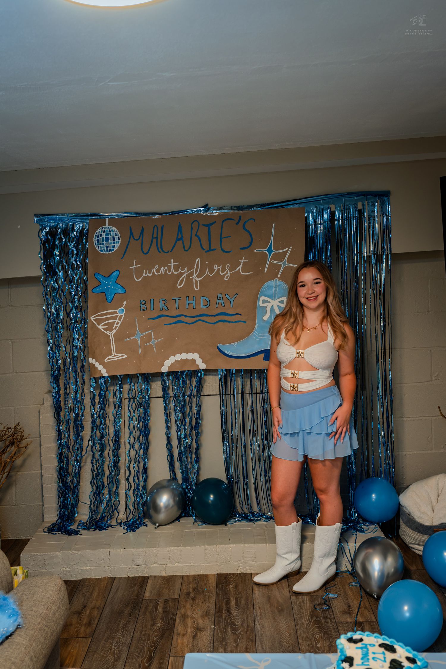 Young woman in blue ruffled skirt and white boots standing before Malarie's 21st birthday party backdrop.