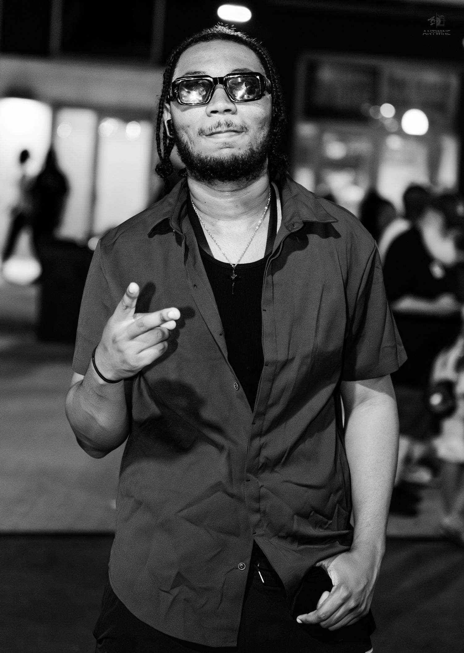 A stylish man in sunglasses and a dark button-up shirt poses with a hand gesture at a nighttime outdoor event.