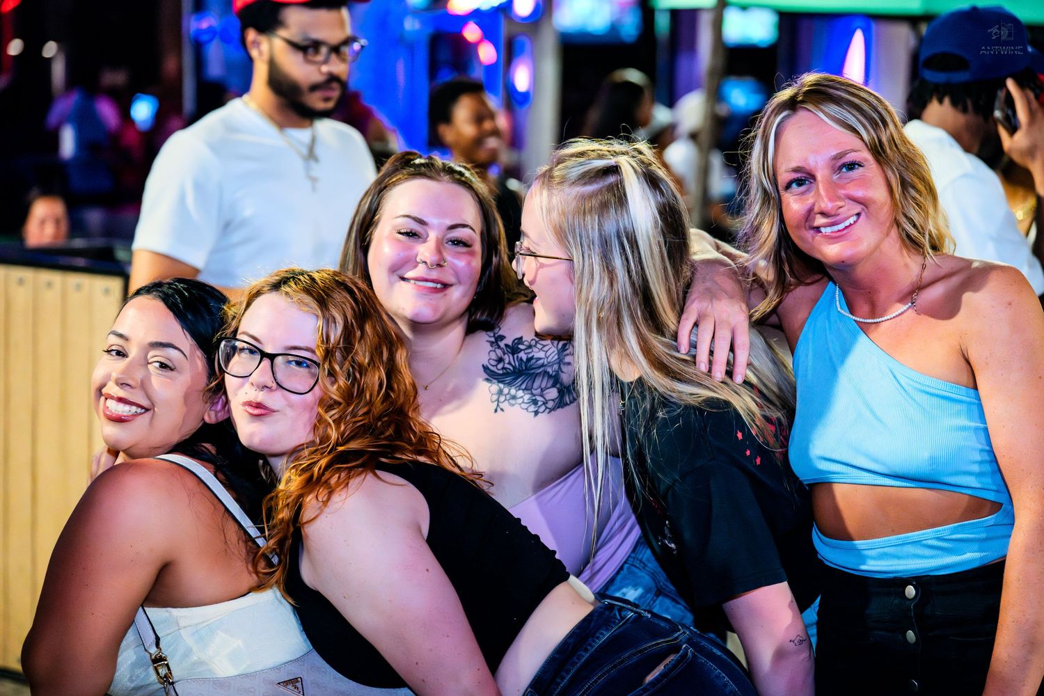 Five young women smiling and posing together at a lively nightlife venue.