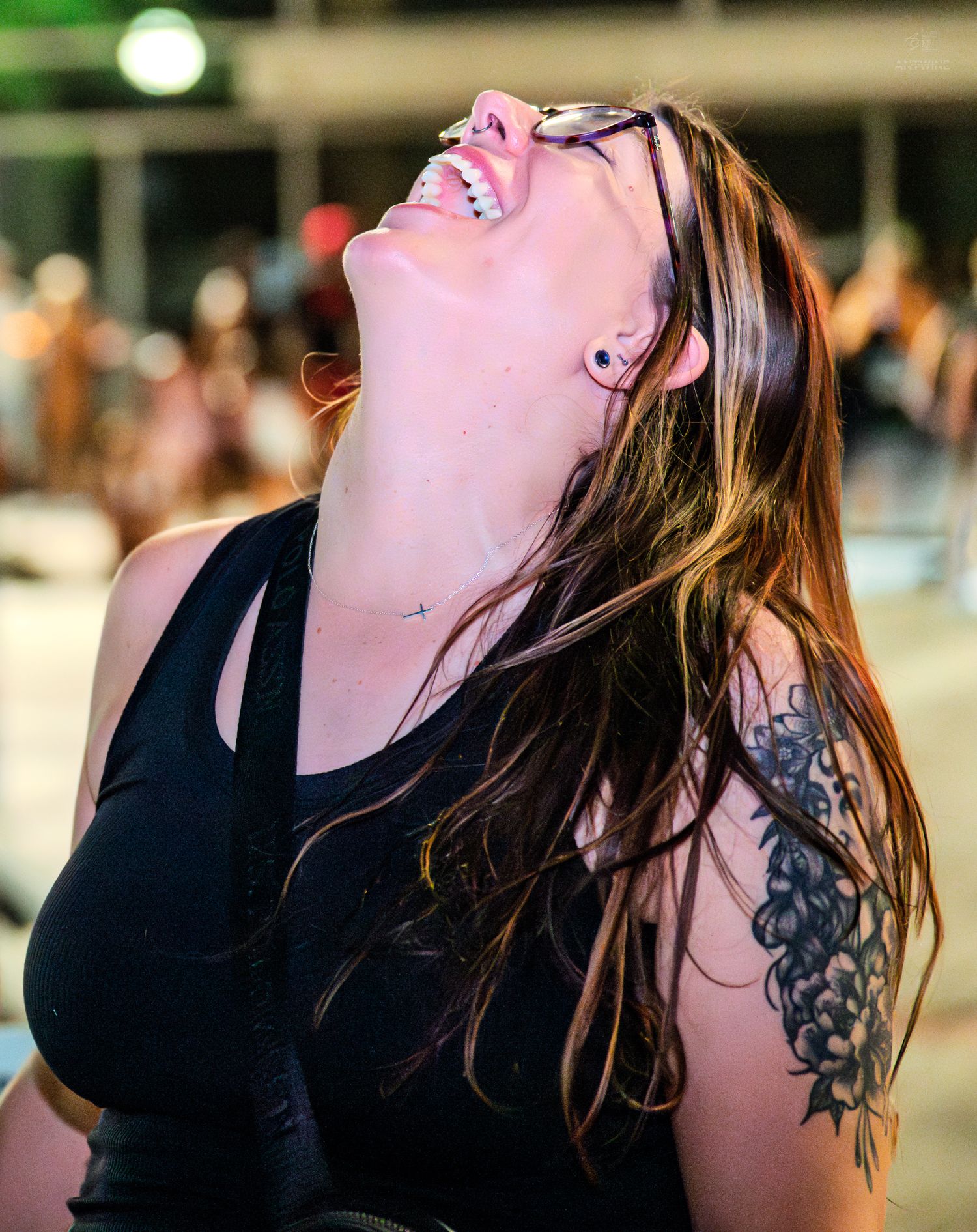 Woman with tattoos and glasses laughing joyfully with head tilted back at outdoor event.