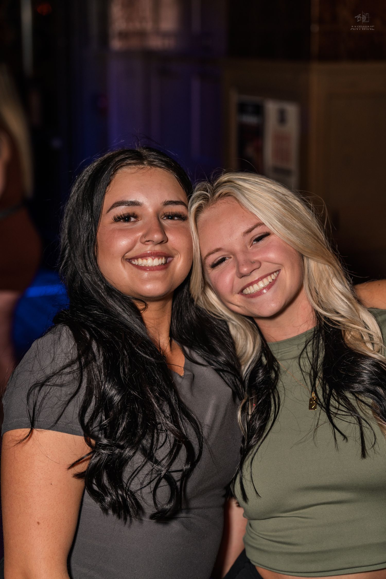 Two smiling young women, one brunette and one blonde, pose together at a bar.