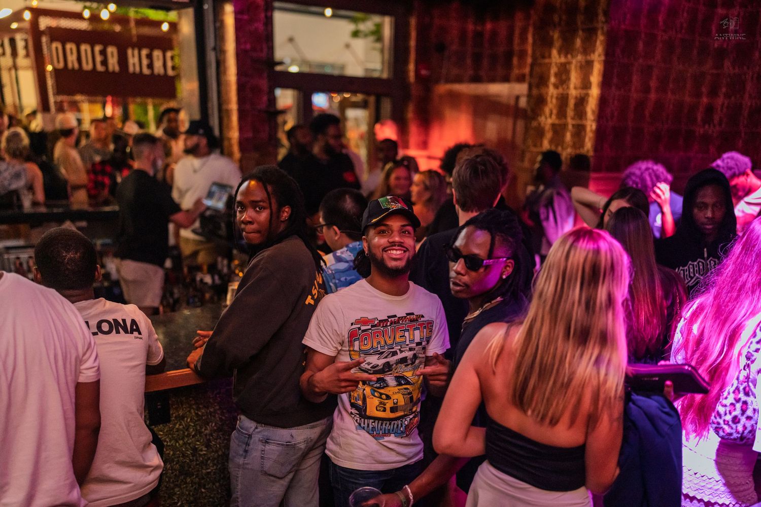 Energetic nightclub crowd with smiling man in graphic tee socializing near an Order Here sign.