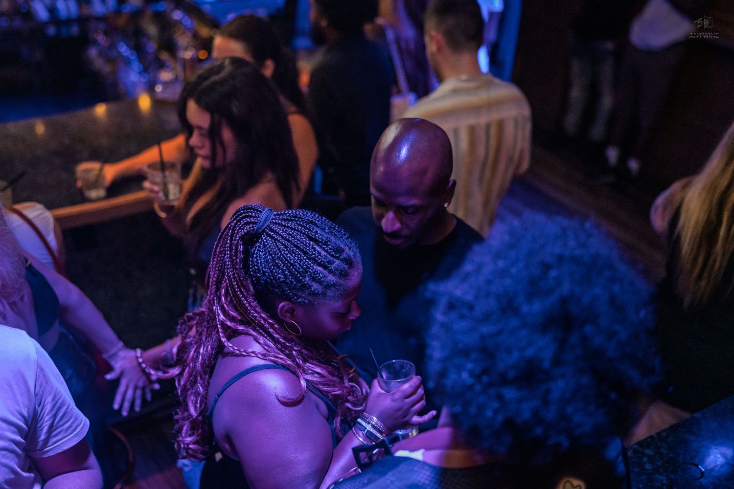 People socializing at a lively nightclub under blue lights, woman with pink braids holding drink.