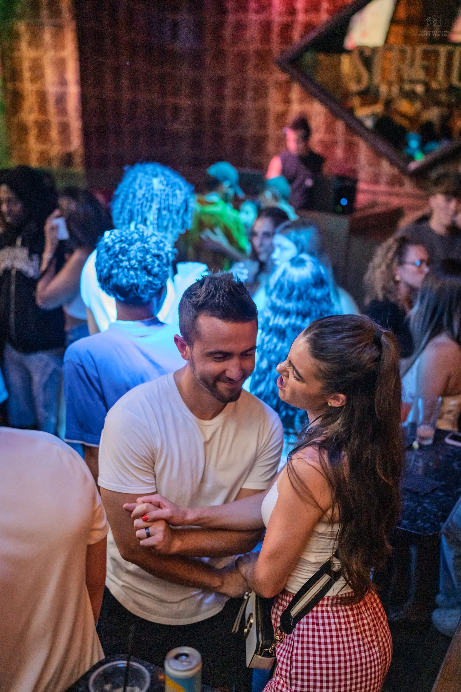 Smiling couple dancing together in a lively nightclub with colorful stage lighting and performers behind them.