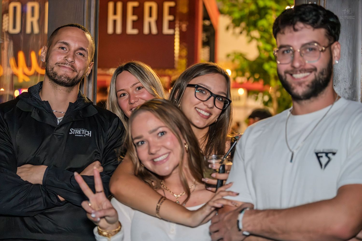 Group of five friends smiling and posing together outside a bar with neon signage.