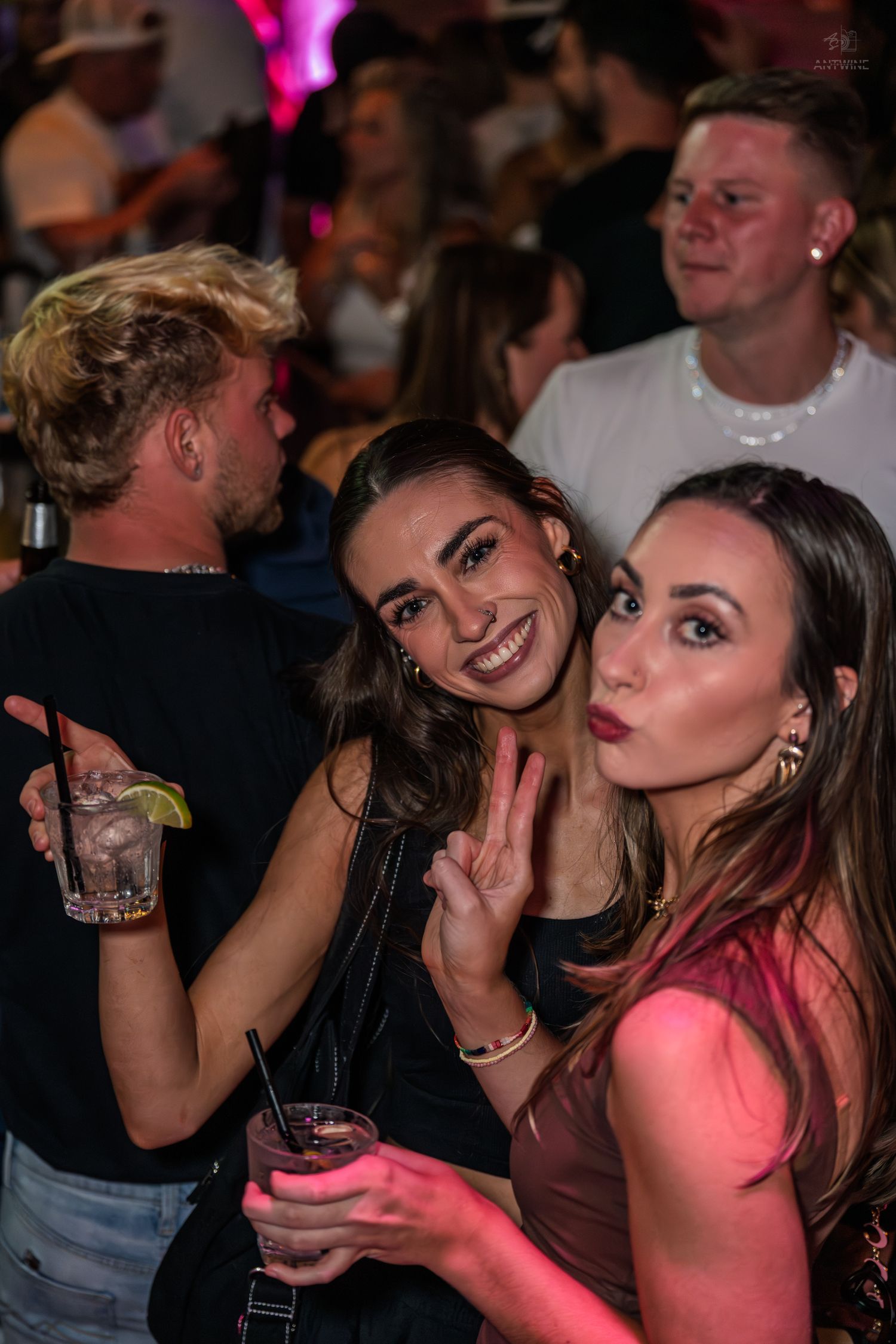 Two smiling young women posing together at a vibrant nightclub party with drinks.