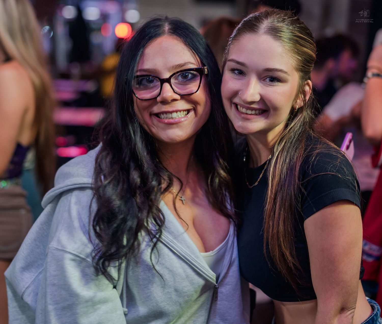 Two smiling women pose together inside a bar, one wearing glasses and gray hoodie, other in black top.