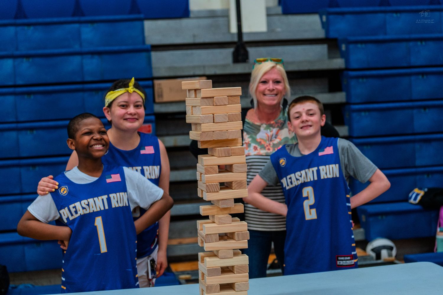 Four people including two Pleasant Run basketball players pose with a giant Jenga tower in a gym.