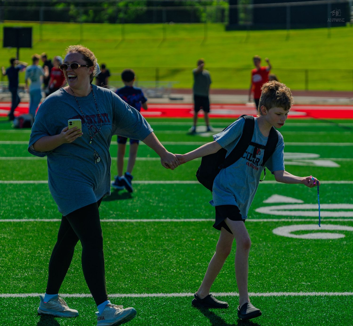 A woman and young boy hold hands while walking on a green turf football field during an outdoor youth sports event.