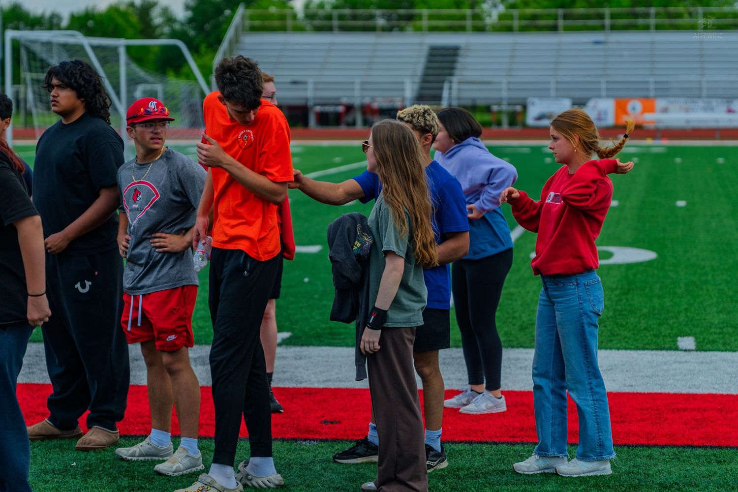 Students line up on a red turf football field during an outdoor school event on a sunny day.