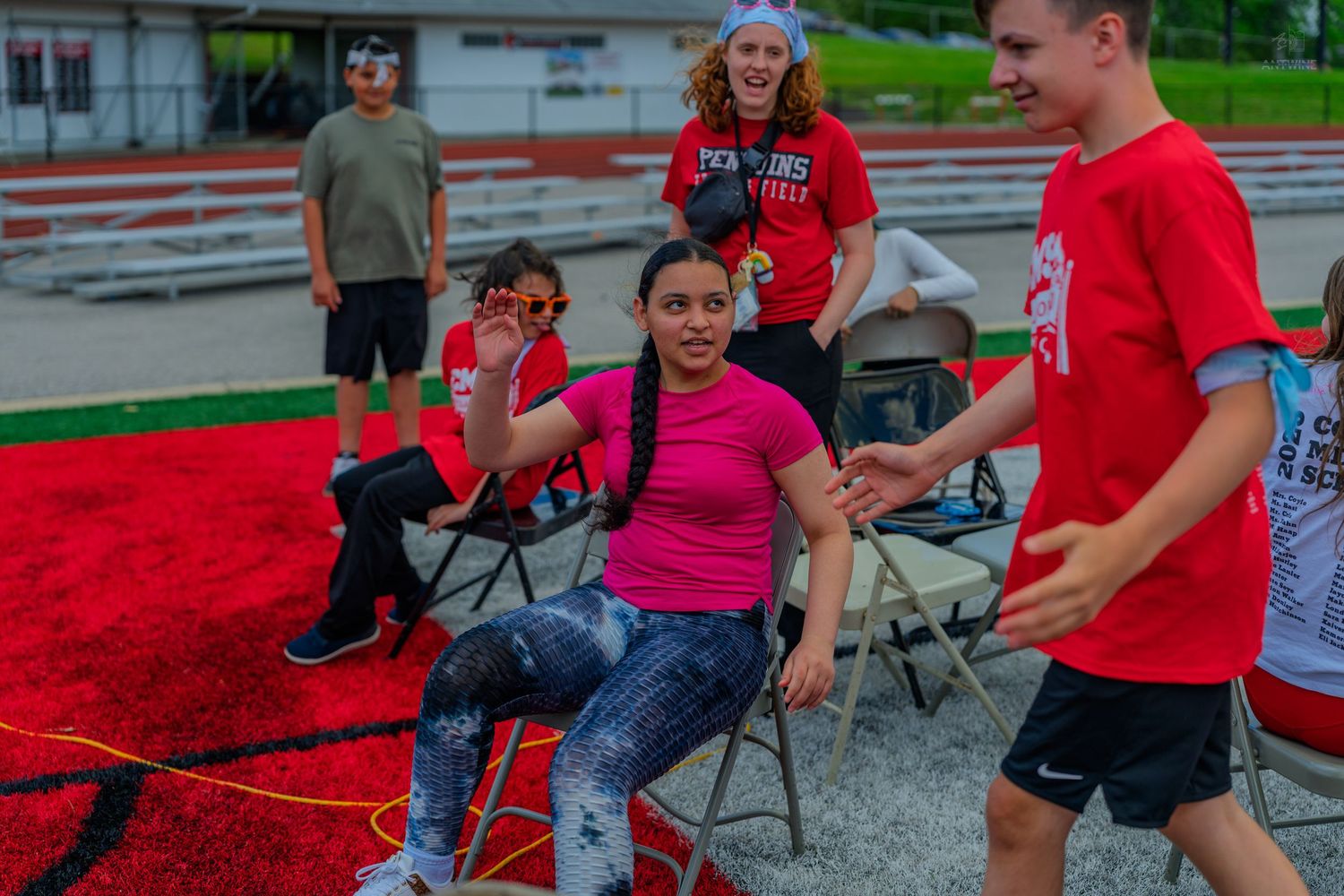 Students in red shirts interact outdoors on a red track field during a school sports event on a summer day.