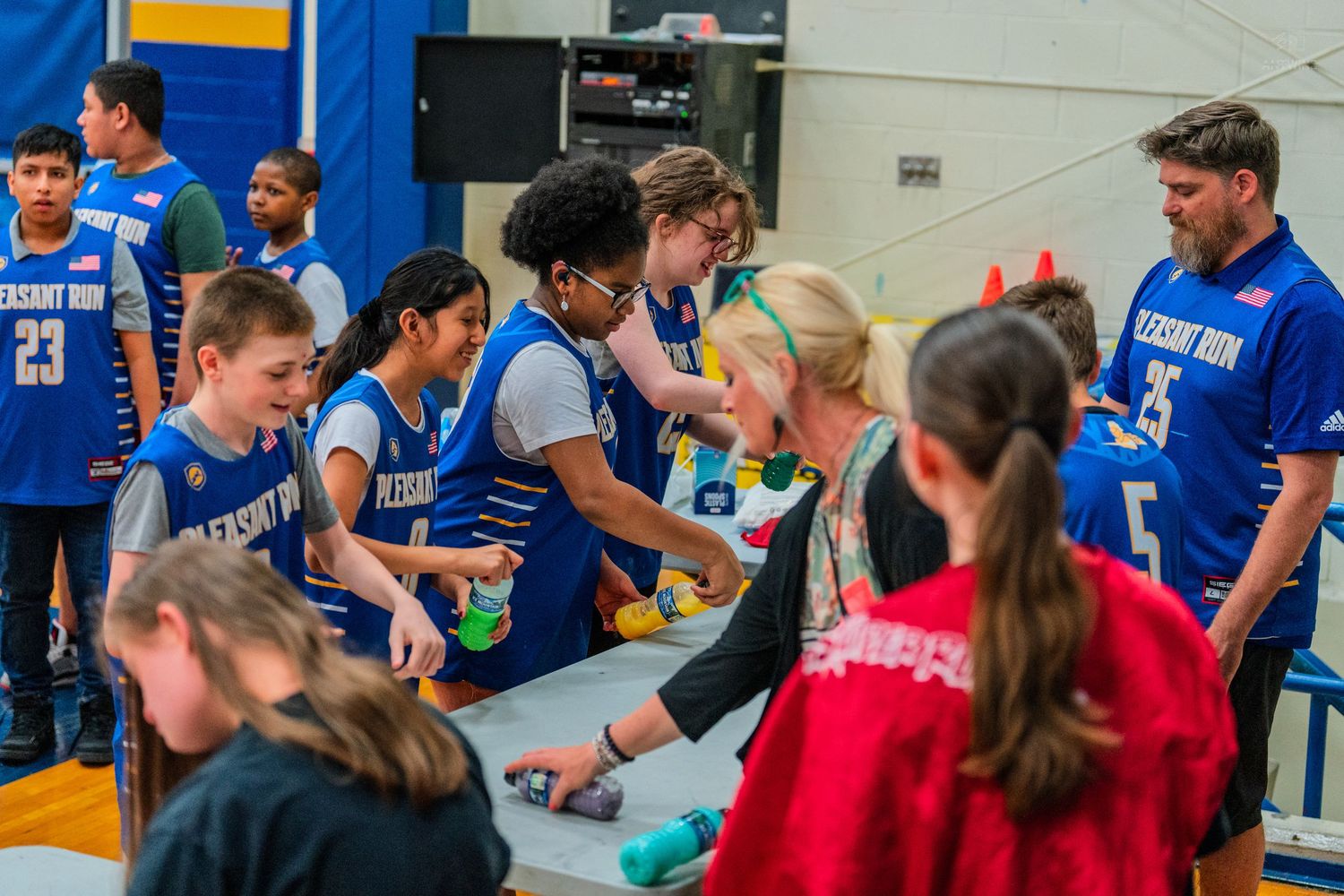 Students in blue jerseys interact with children at a table during an indoor basketball gym event.