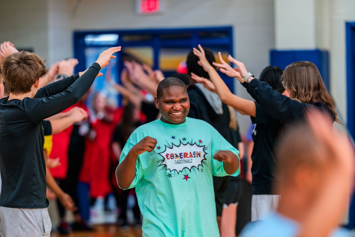 Student in green Comerica shirt smiles while peers raise hands in celebration during school gym activity.