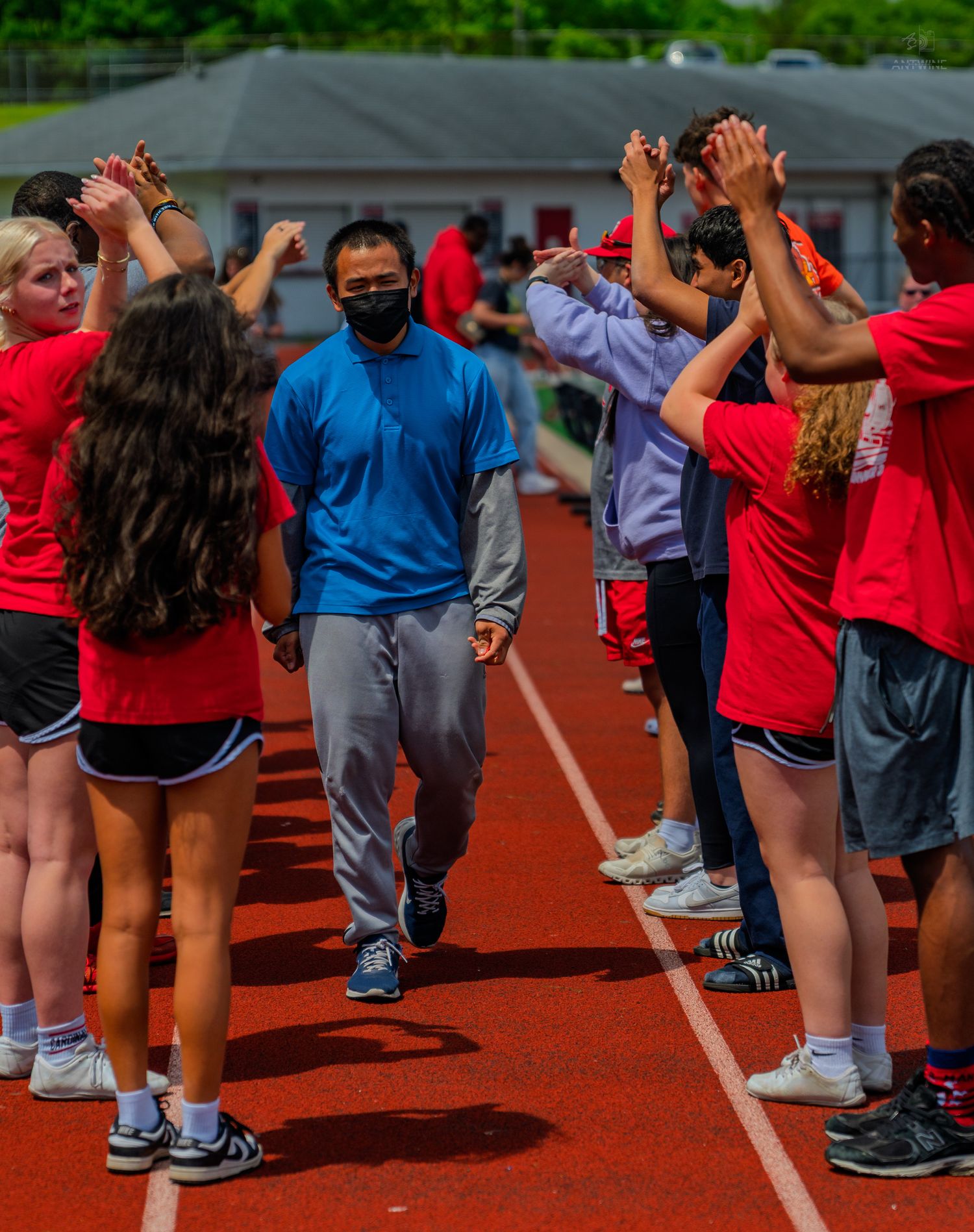 Students in red shirts form a congratulatory tunnel on a track, high-fiving a masked athlete in blue.