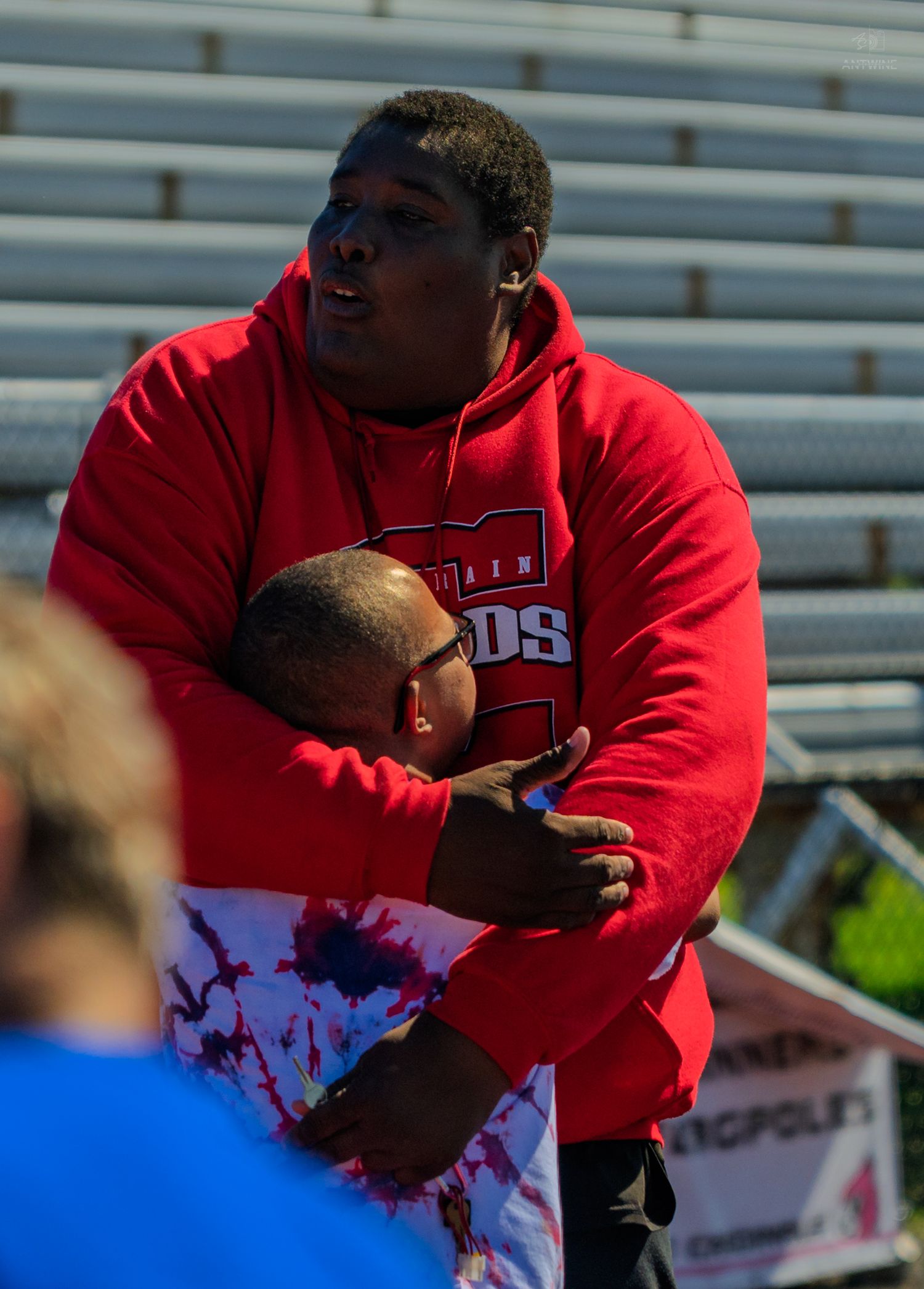 Two people in red hoodies sharing a warm hug at an outdoor sports stadium event.