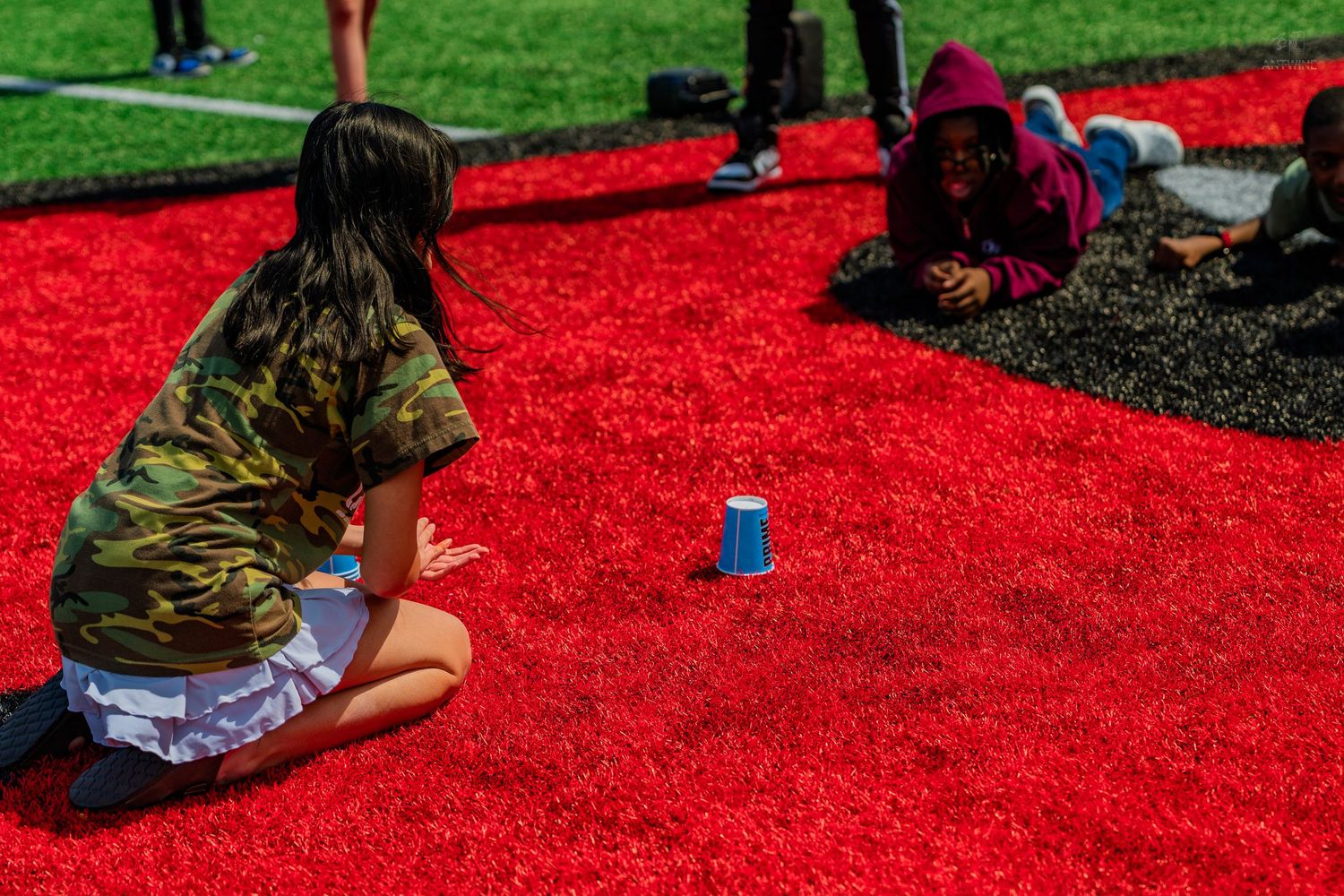 Two children playing a cup game on bright red artificial turf field on a sunny day.