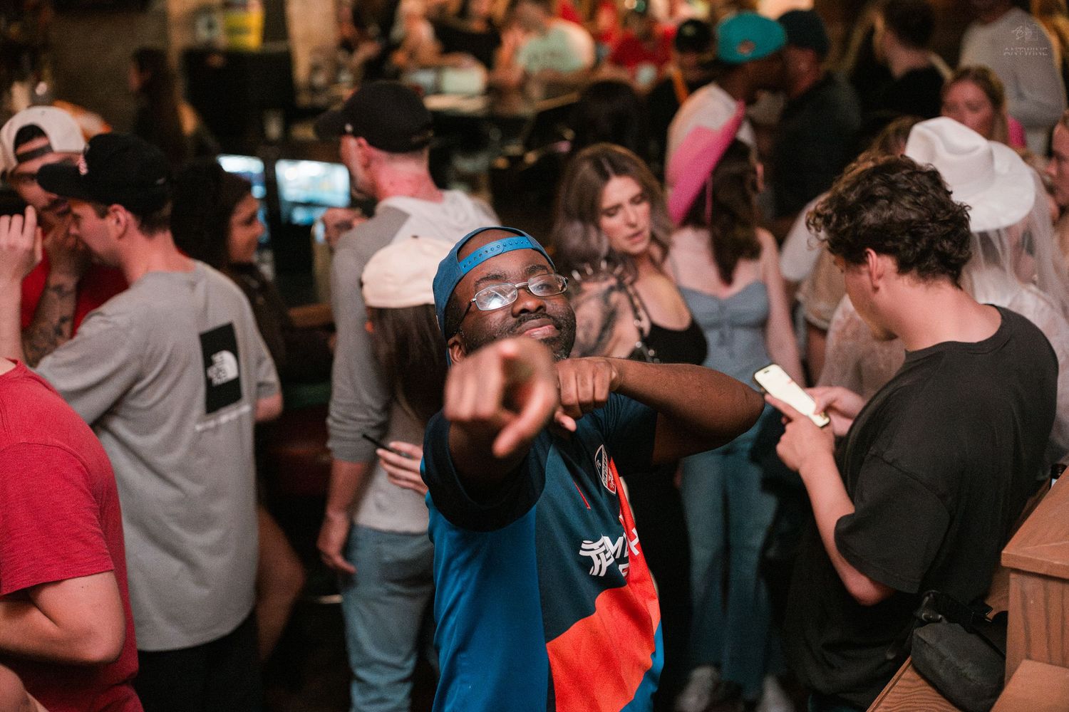 A lively man in a blue hoodie points at the camera while dancing at a crowded Cincinnati bar party.