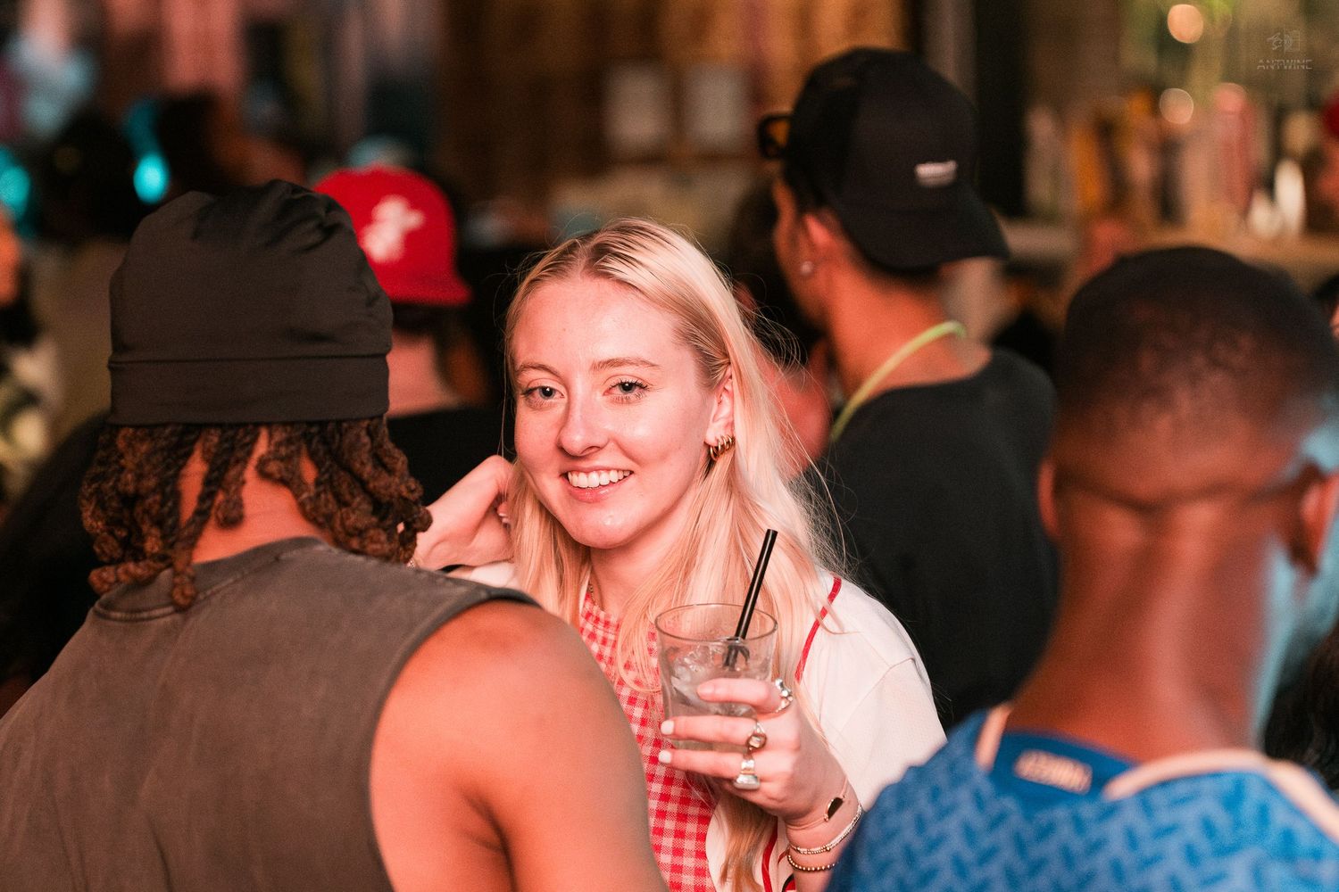 Smiling blonde woman holding a drink at a crowded bar or nightclub event.