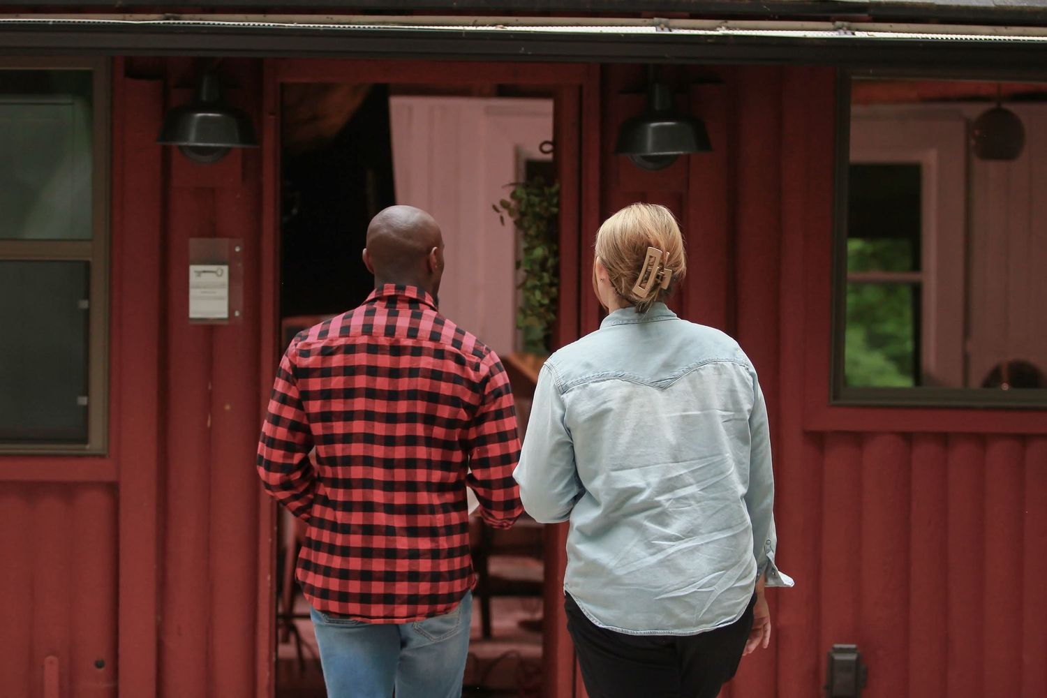 Two people holding hands walking toward a red building, man in plaid shirt and woman in light blue jacket.