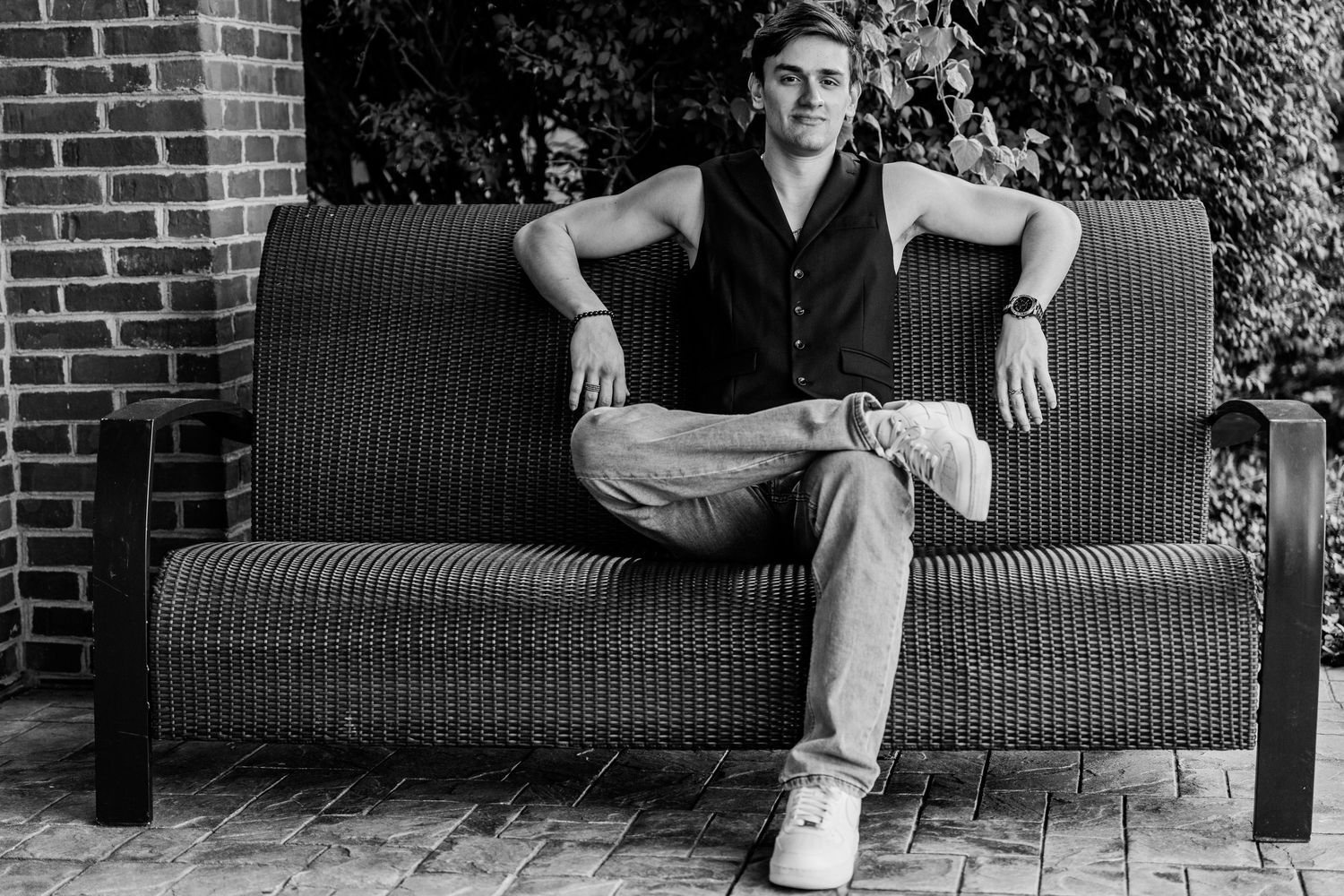 Young man in black vest and jeans sitting relaxed on outdoor wicker sofa, black and white portrait photo.