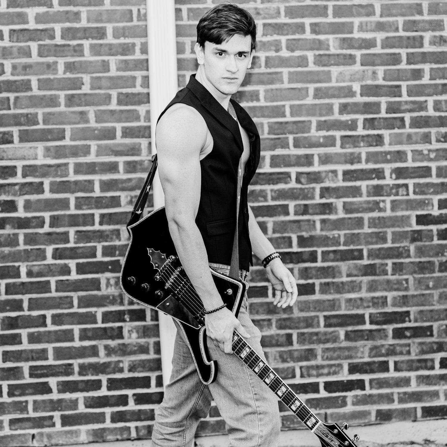 Young male musician in black sleeveless vest carrying an electric guitar while walking past a brick wall.