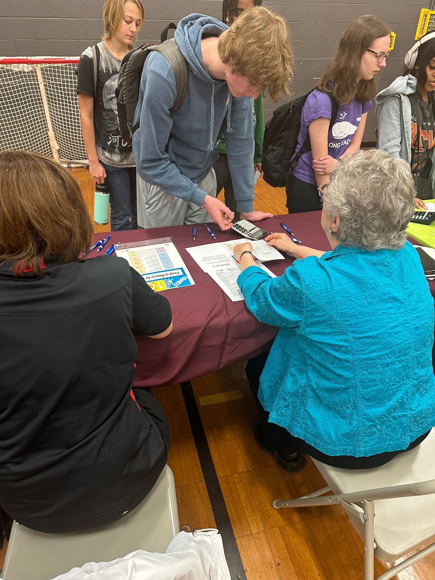 Students interact with elderly volunteer at informational table during school gymnasium event or fair.