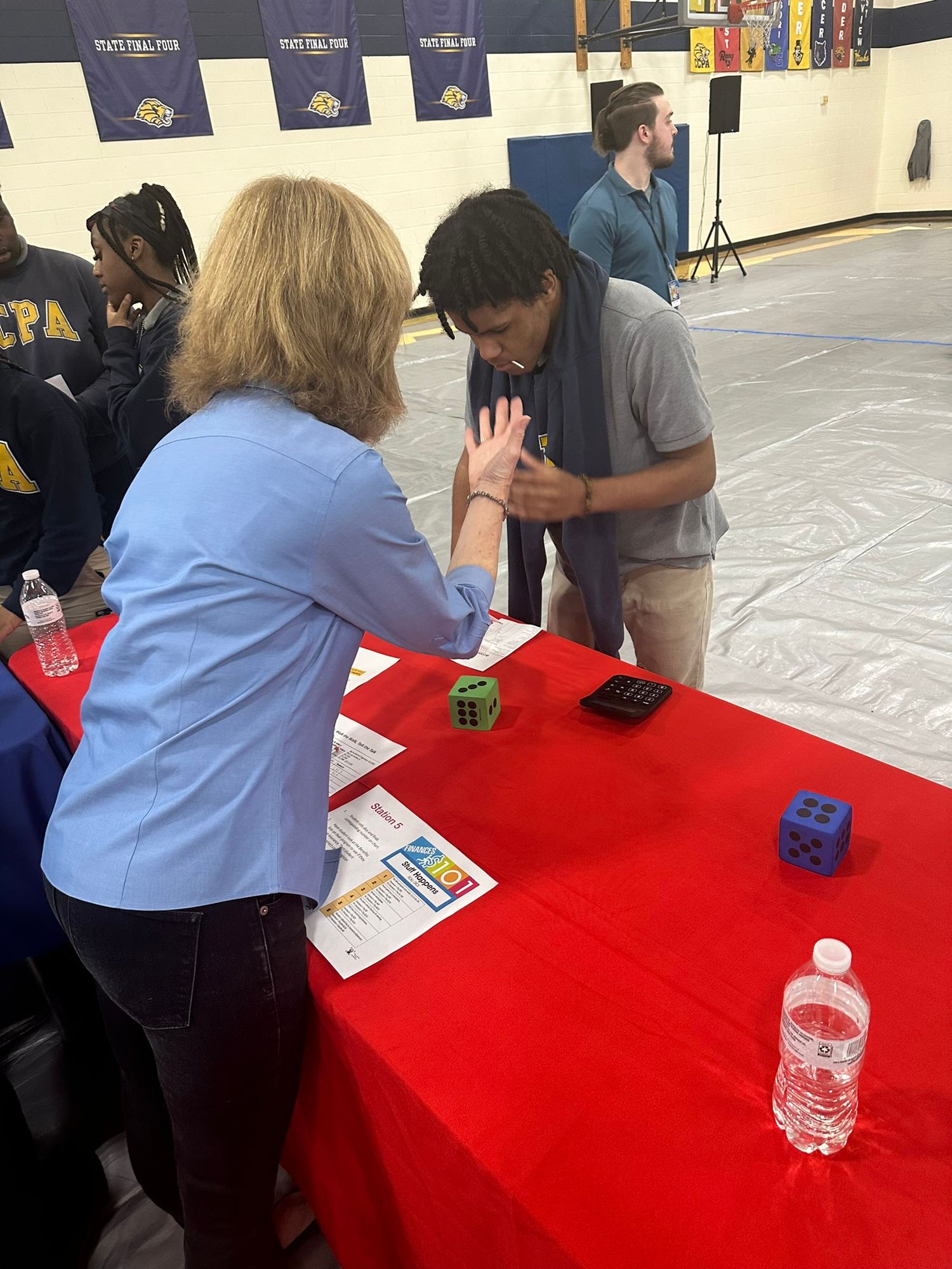 A woman and student interact over dice and calculators at a red table during a school financial literacy fair.