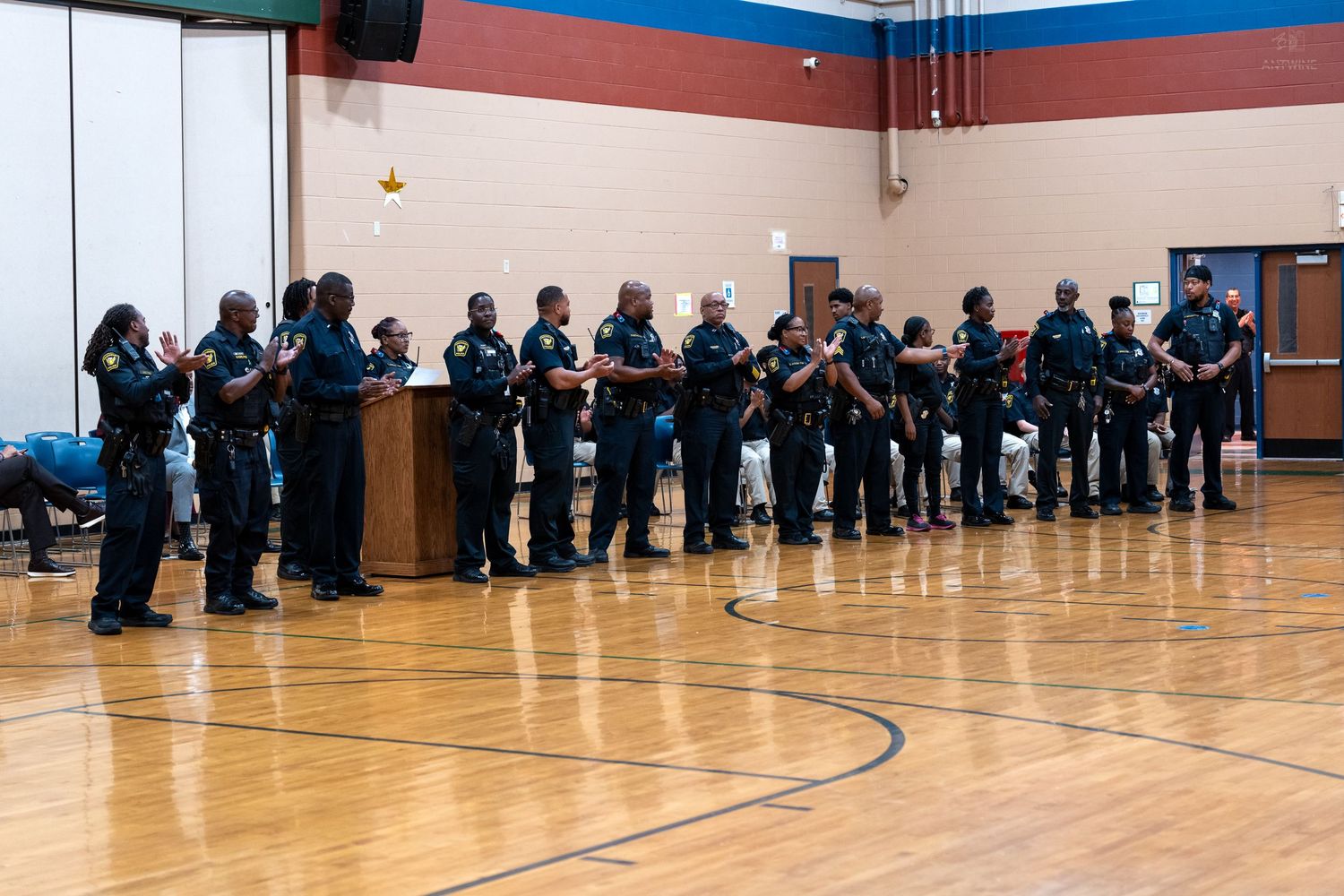 Police officers stand in a line during an indoor ceremony in a gymnasium, wearing uniforms with badges and equipment.