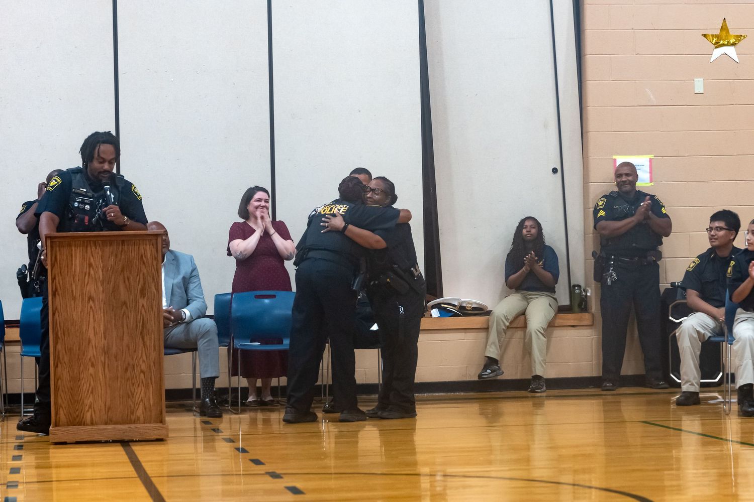Two people share a heartfelt hug on a gym stage during a police recognition ceremony while audience members applaud.
